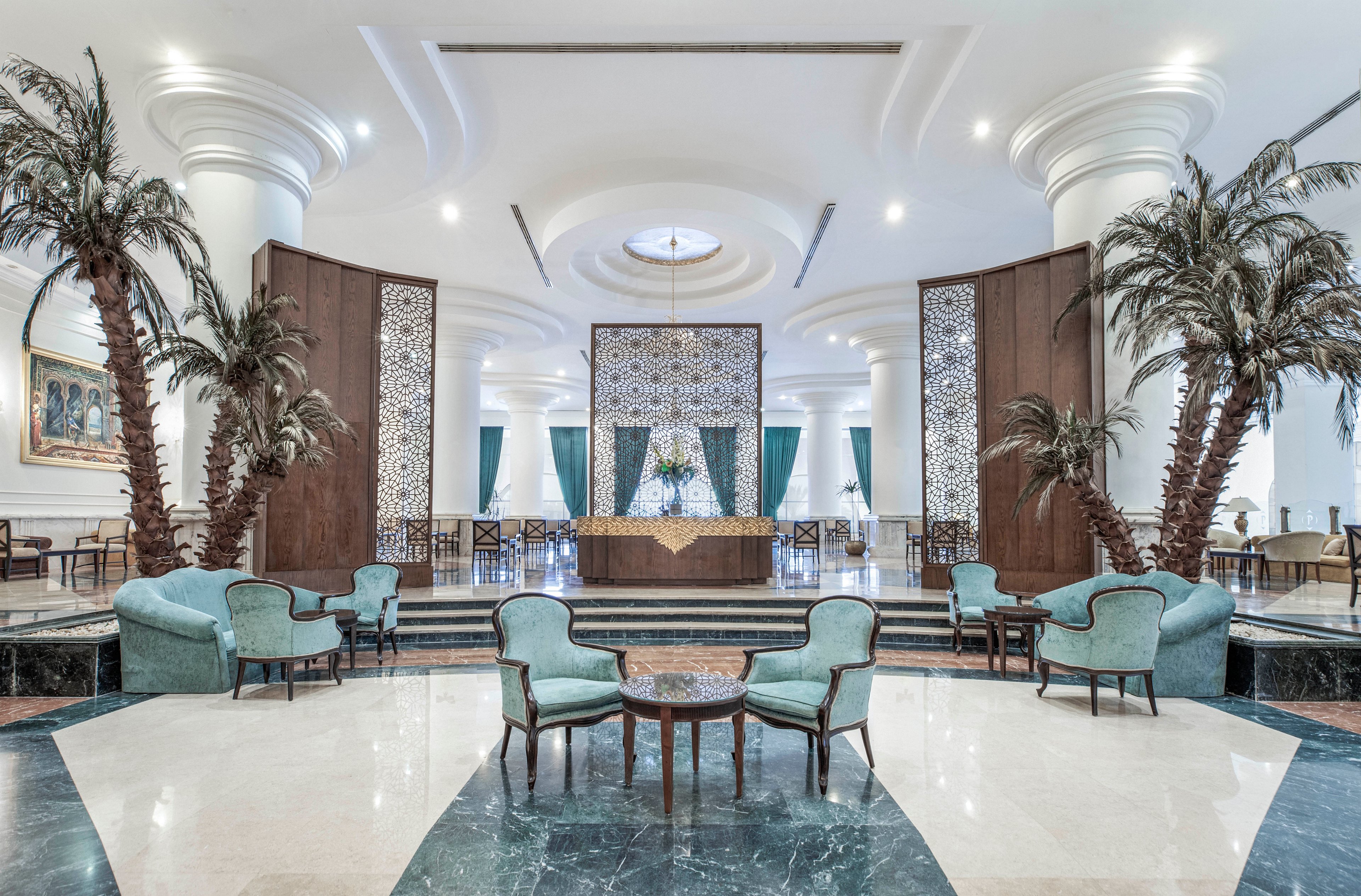 A spacious hotel lobby features high white ceilings, large columns, and decorative palm trees. Vintage-style armchairs and tables are arranged on a polished marble floor, with patterned screens and draped curtains in the background. The setting is bright, airy, and sophisticated, with a cool color palette and symmetrical design.