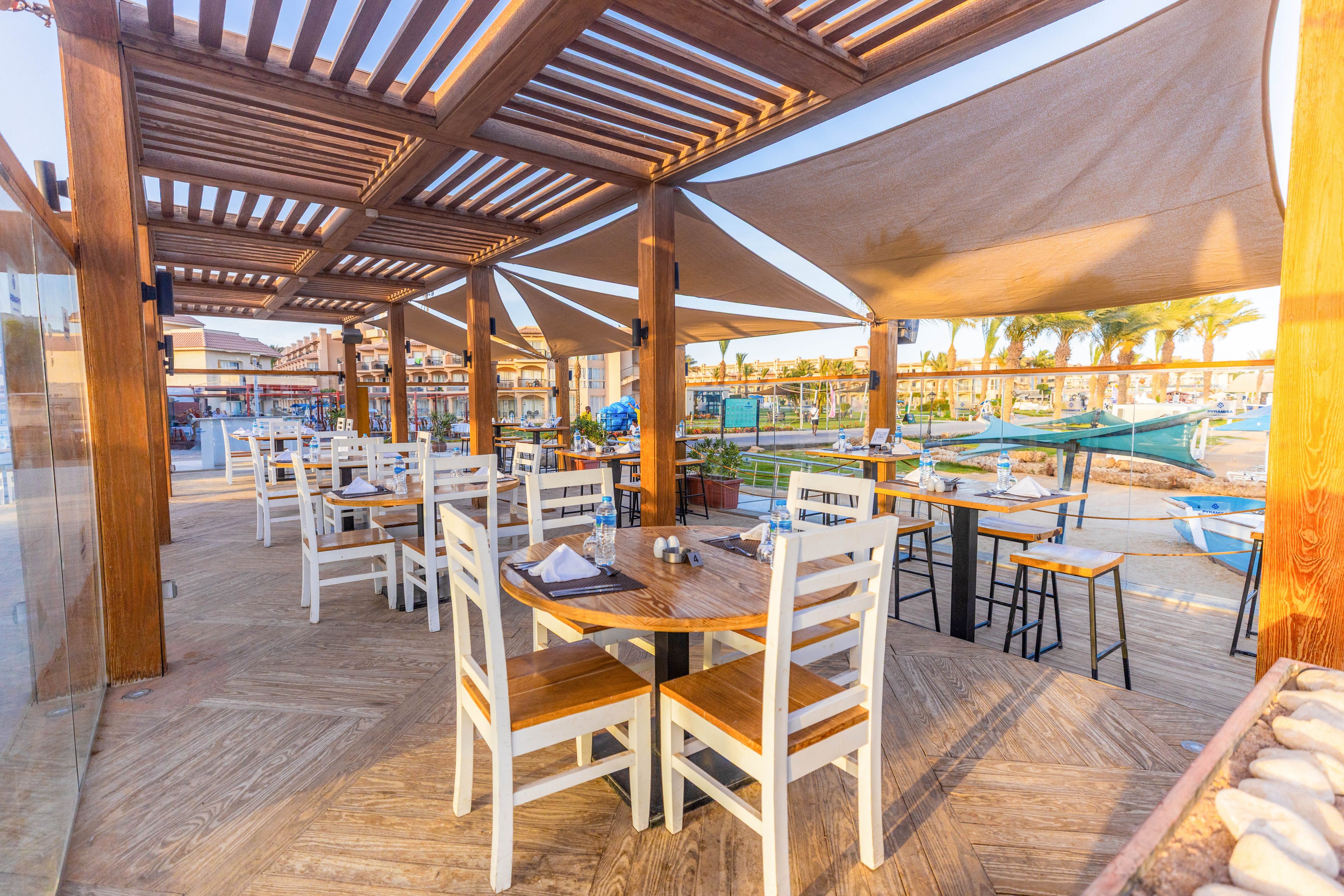 An open-air restaurant patio features wooden pergolas and neatly arranged tables with white chairs. The setting is bright and sunny, with palm trees and a pool visible in the background, suggesting a resort or vacation atmosphere. The area is empty, emphasizing a tranquil and inviting mood with natural wood tones and blue accents.