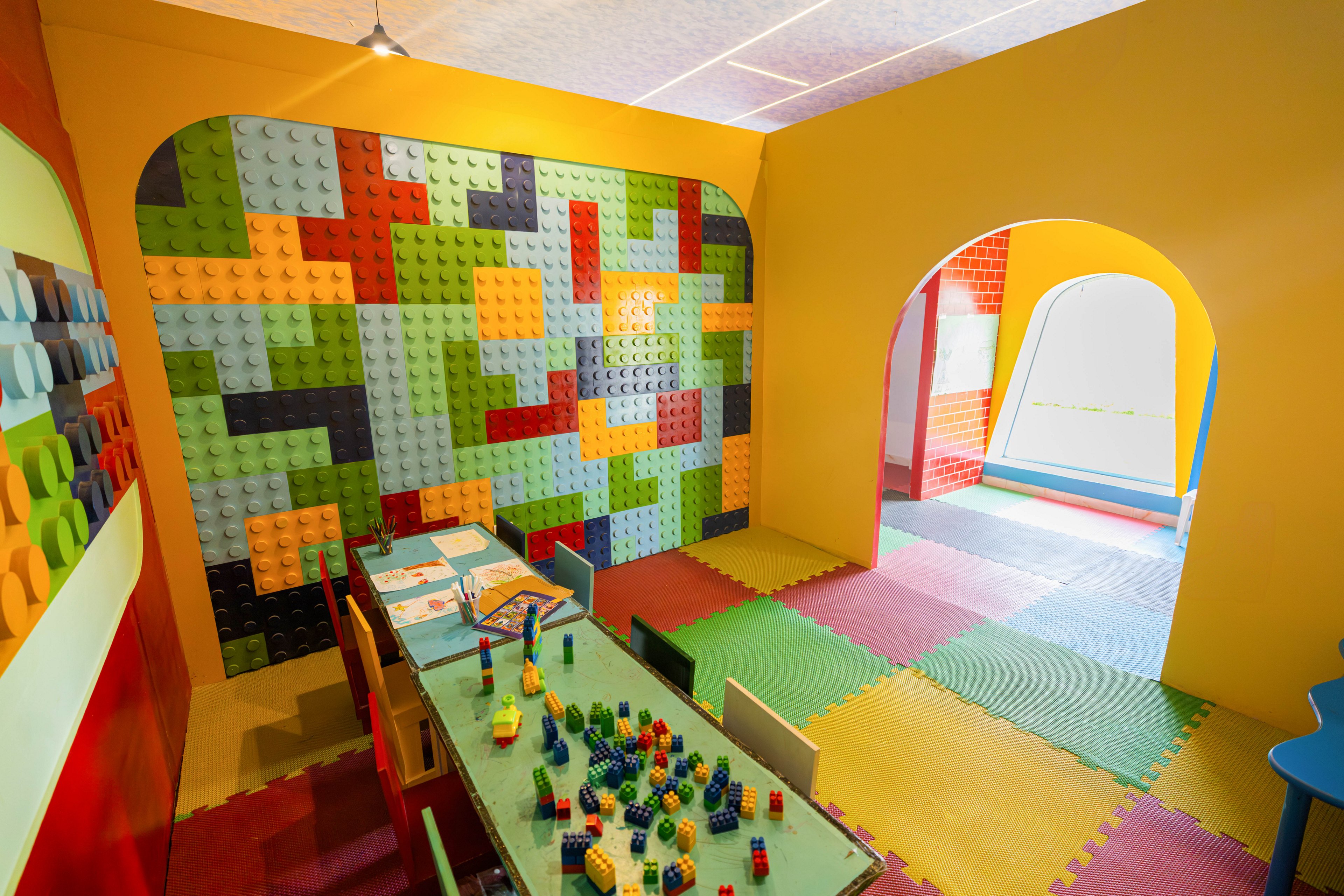 A vibrant children's playroom features a wall decorated with large, colorful interlocking blocks. The room includes a table with various building blocks and chairs, set on a floor covered with multicolored foam mats. Bright yellow and red walls, along with arched doorways, create a playful and inviting atmosphere. The space is designed for creative play and group activities.