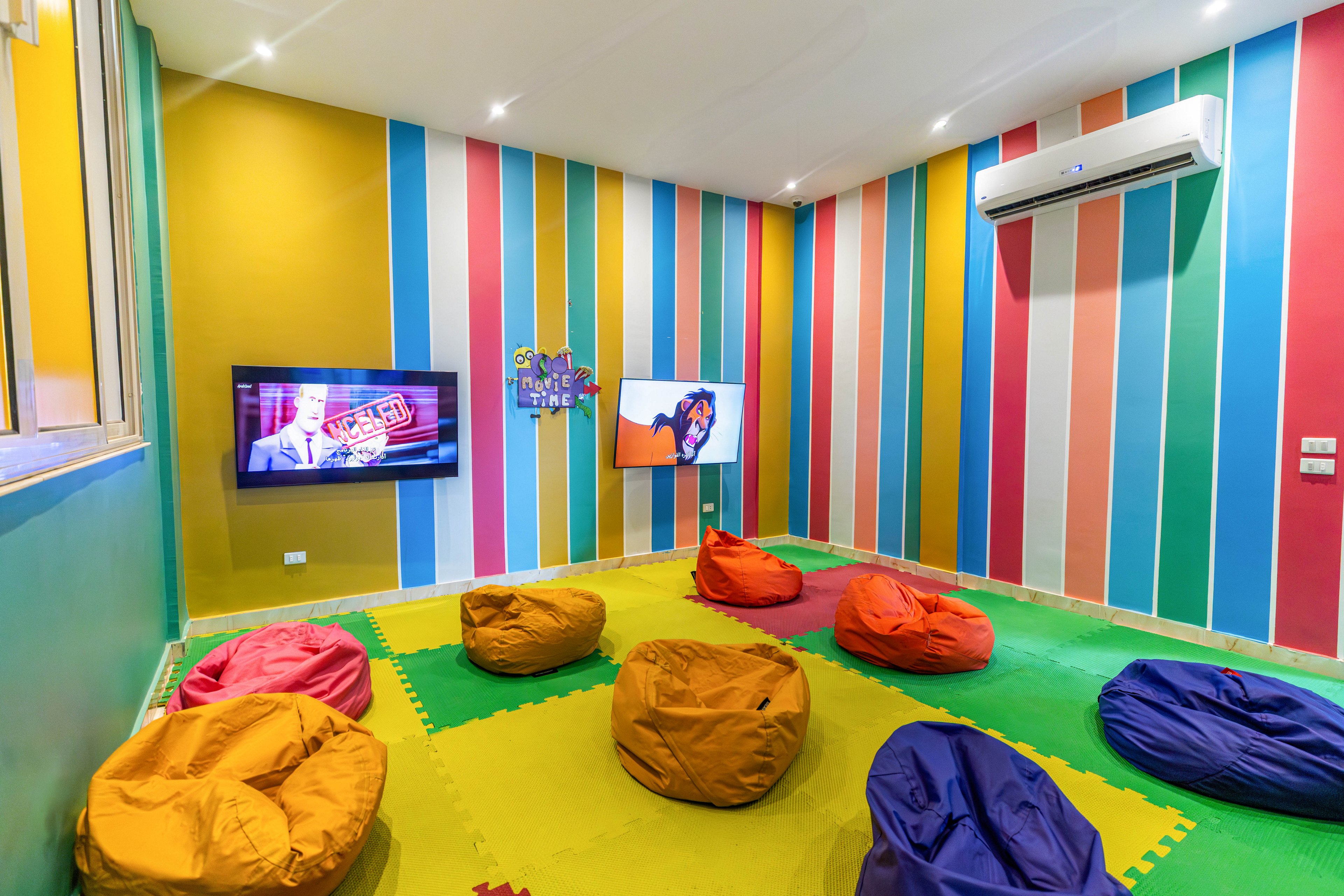 A vibrant room features multicolored vertical striped walls and a soft foam floor with interlocking mats. Several bean bag chairs in yellow, orange, red, and purple are arranged for casual seating. Two wall-mounted televisions display different content, creating a playful and relaxed atmosphere. The space is brightly lit and equipped with an air conditioning unit.