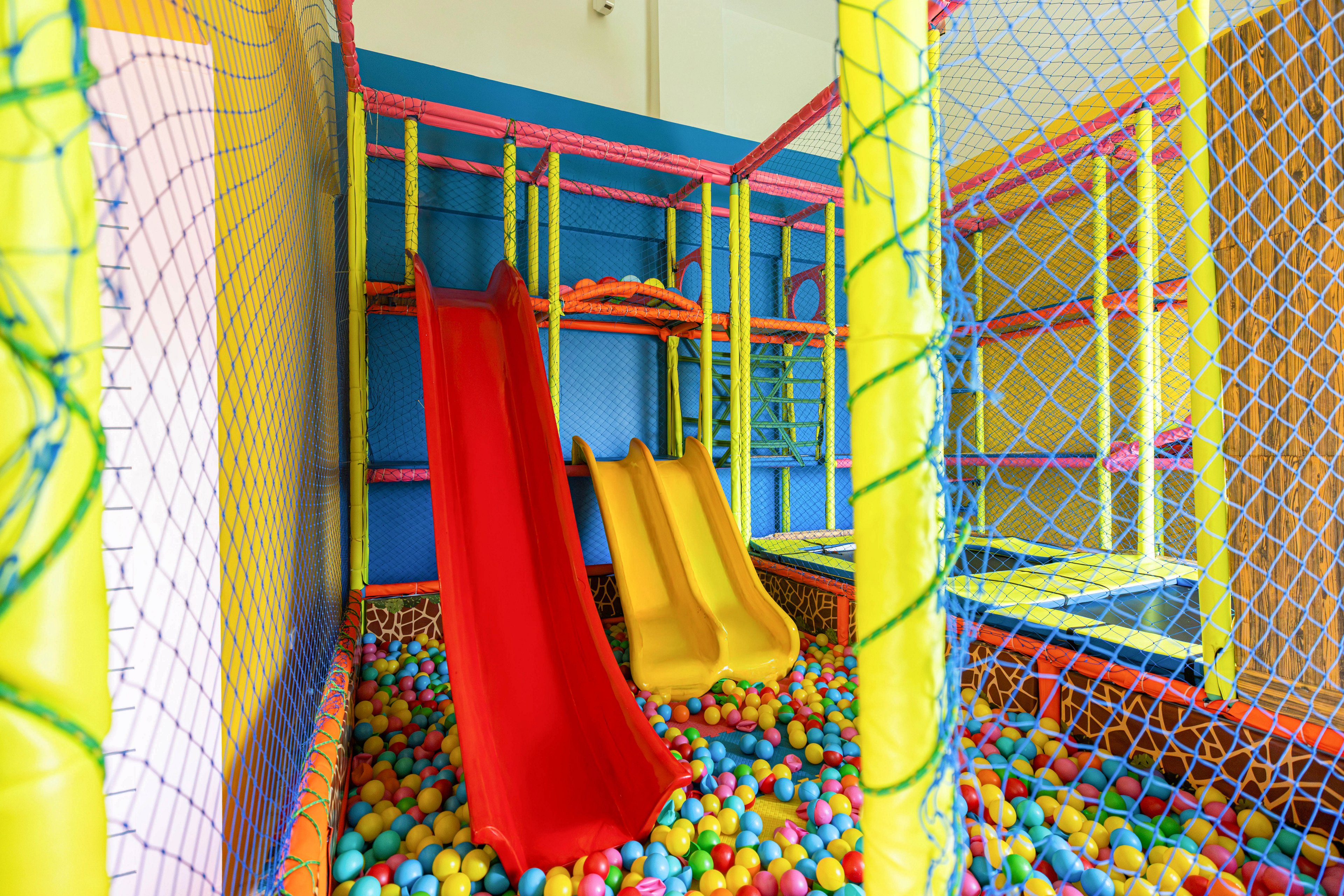 A vibrant indoor playground features two slides, one red and one yellow, leading into a multicolored ball pit. The play area is enclosed with safety netting and surrounded by padded structures in bright primary colors. The setting is designed for children, offering a fun and safe environment for active play. No visible text or numbers are present in the image.