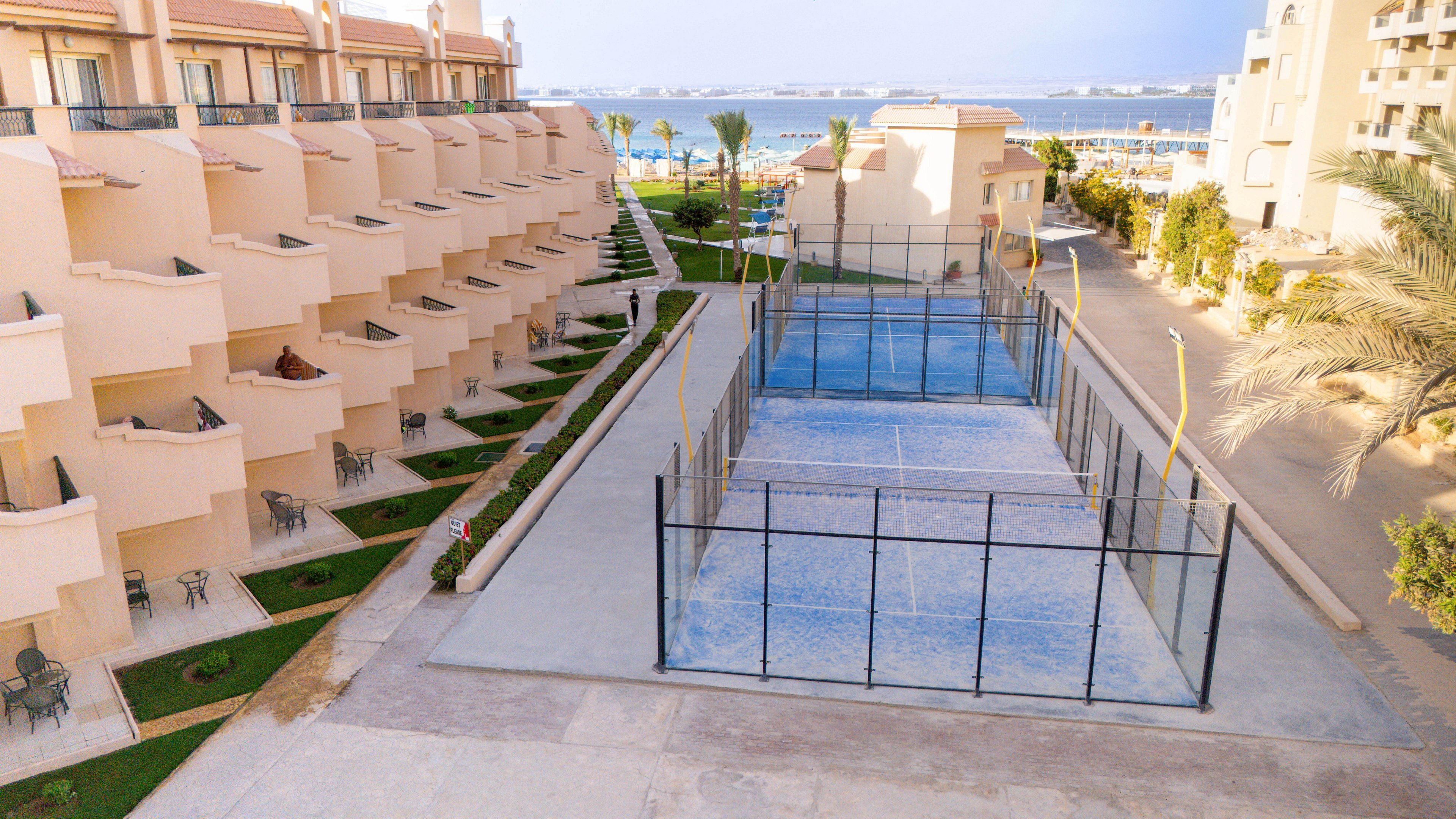 Aerial view of a contemporary resort complex featuring multiple balconies overlooking a fenced outdoor sports court. The setting is bright and sunny, with palm trees and landscaped pathways leading toward a blue sea in the background. The architecture is light beige, and the overall mood is relaxed and inviting, ideal for leisure and recreation.