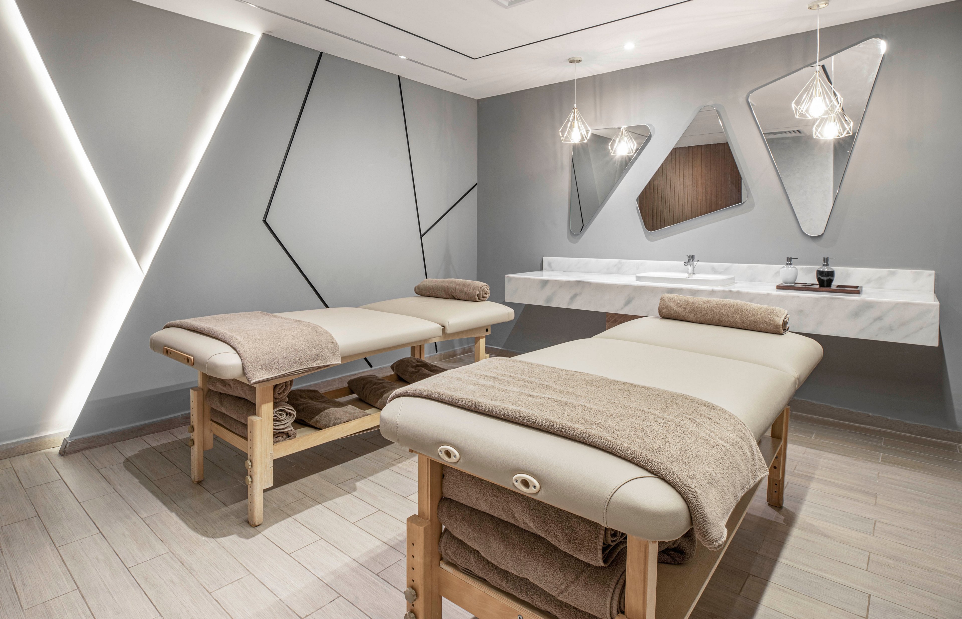 A contemporary spa room features two padded massage tables with rolled towels, set against a backdrop of geometric wall designs and soft lighting. The space includes a sleek marble countertop with two sinks and decorative mirrors. The neutral color palette and minimalist decor create a calm, relaxing atmosphere ideal for wellness treatments.