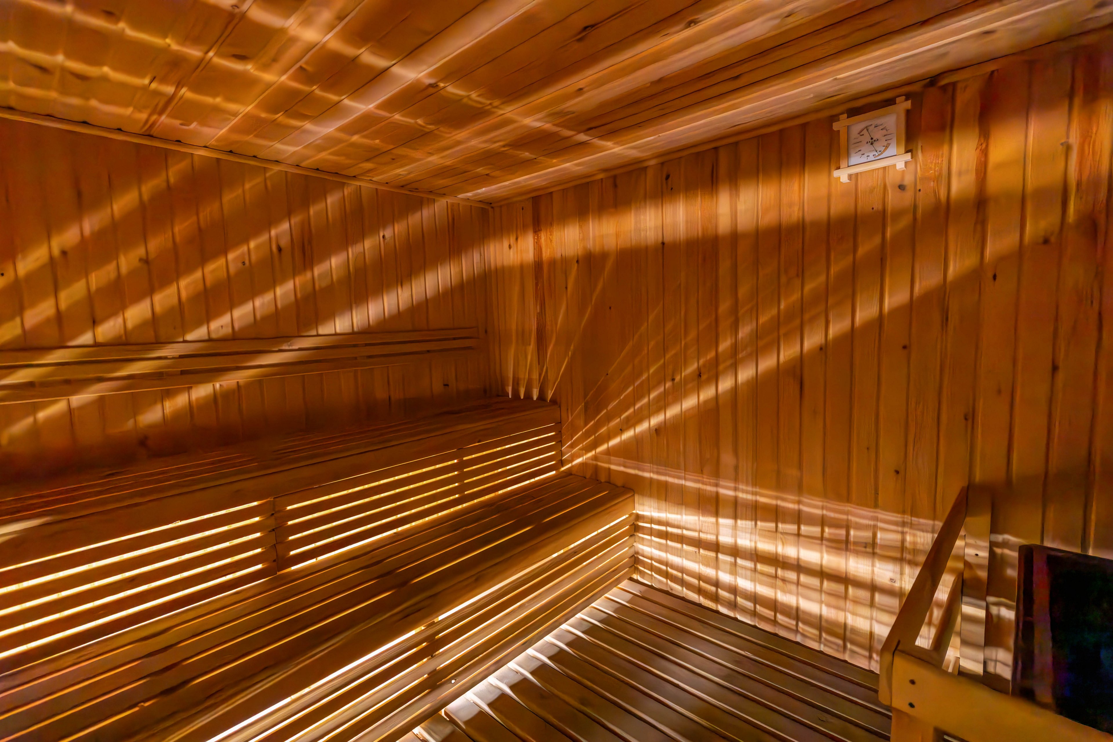 A spacious wooden sauna room is illuminated by sunlight streaming through slats, casting dramatic patterns on the walls and benches. The interior features tiered seating and a warm, inviting wood finish. No people are present, emphasizing the tranquil and serene atmosphere. The overall mood is calm and relaxing, with a natural color palette dominated by warm wood tones.