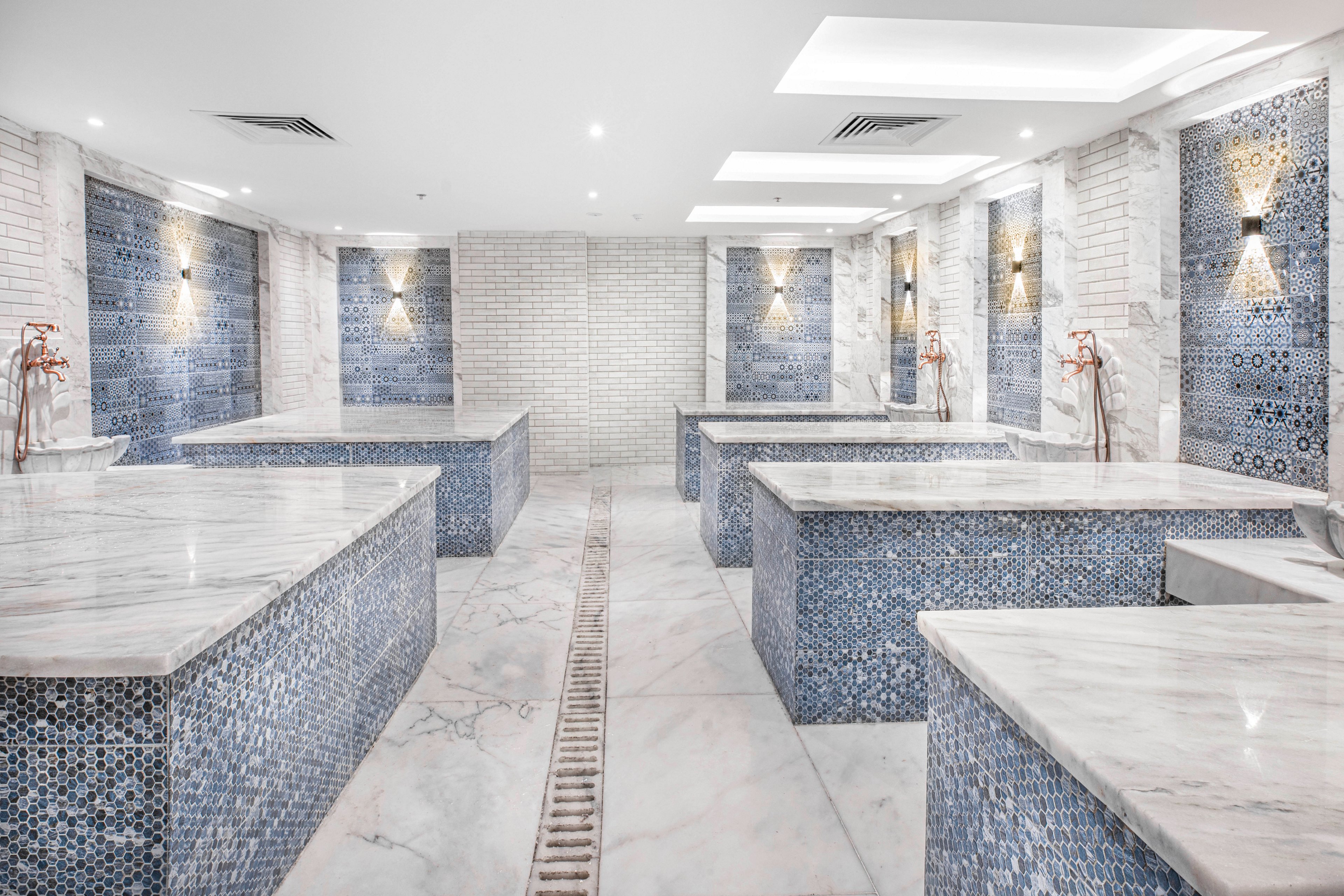 A spacious Turkish hammam spa features multiple marble massage tables with intricate blue mosaic tile bases. The setting is bright and clean, with white brick walls and recessed lighting creating a serene atmosphere. Decorative wall sconces add warmth, while the overall palette is cool and calming. No people are present, emphasizing the tranquil and inviting environment.