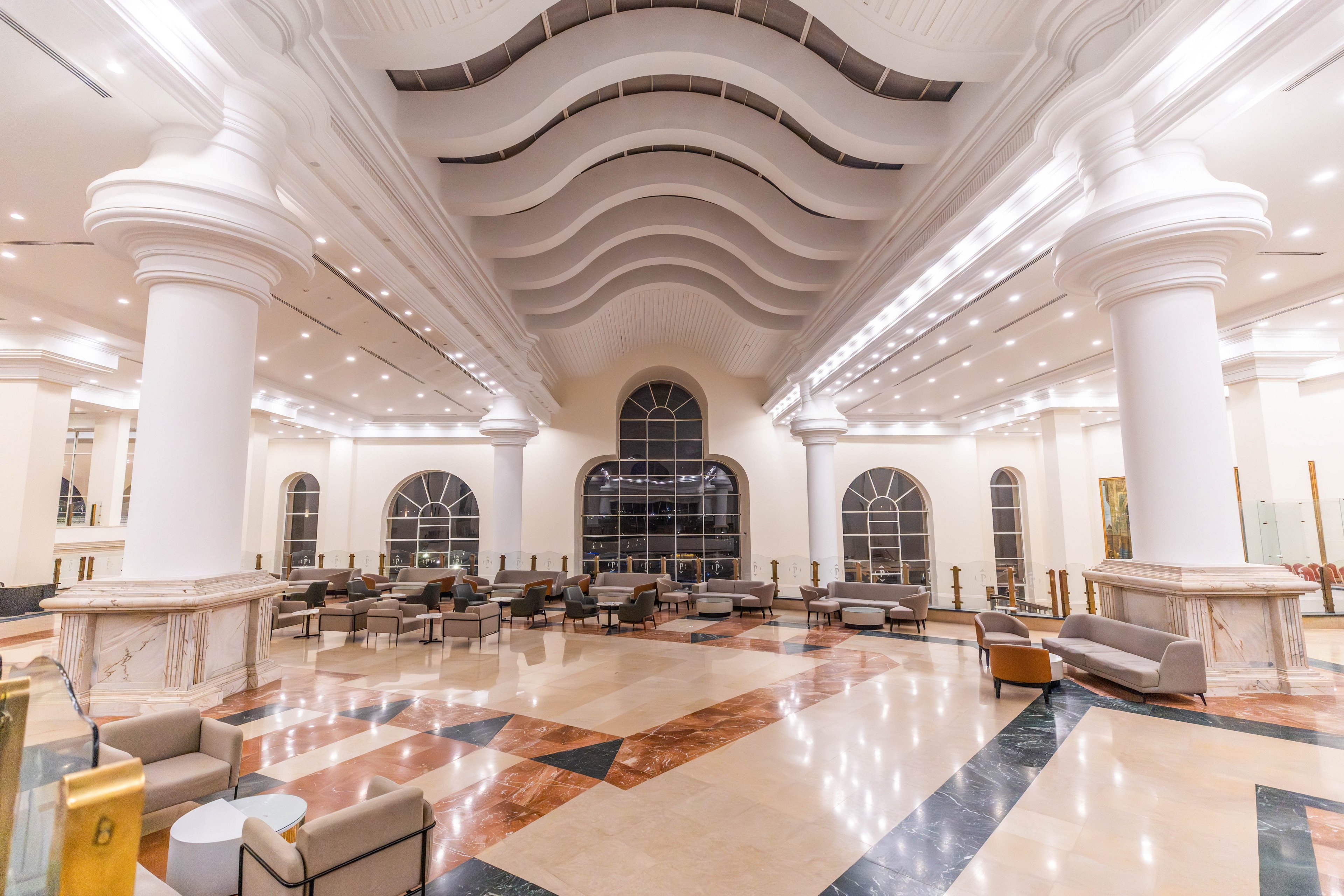 A grand hotel lobby features high ceilings with wave-like architectural details and large arched windows. The space is illuminated by bright overhead lighting, highlighting marble floors with geometric patterns. Comfortable armchairs and sofas are arranged throughout the open area, creating a welcoming and luxurious atmosphere. The setting is empty, emphasizing the clean and serene environment.