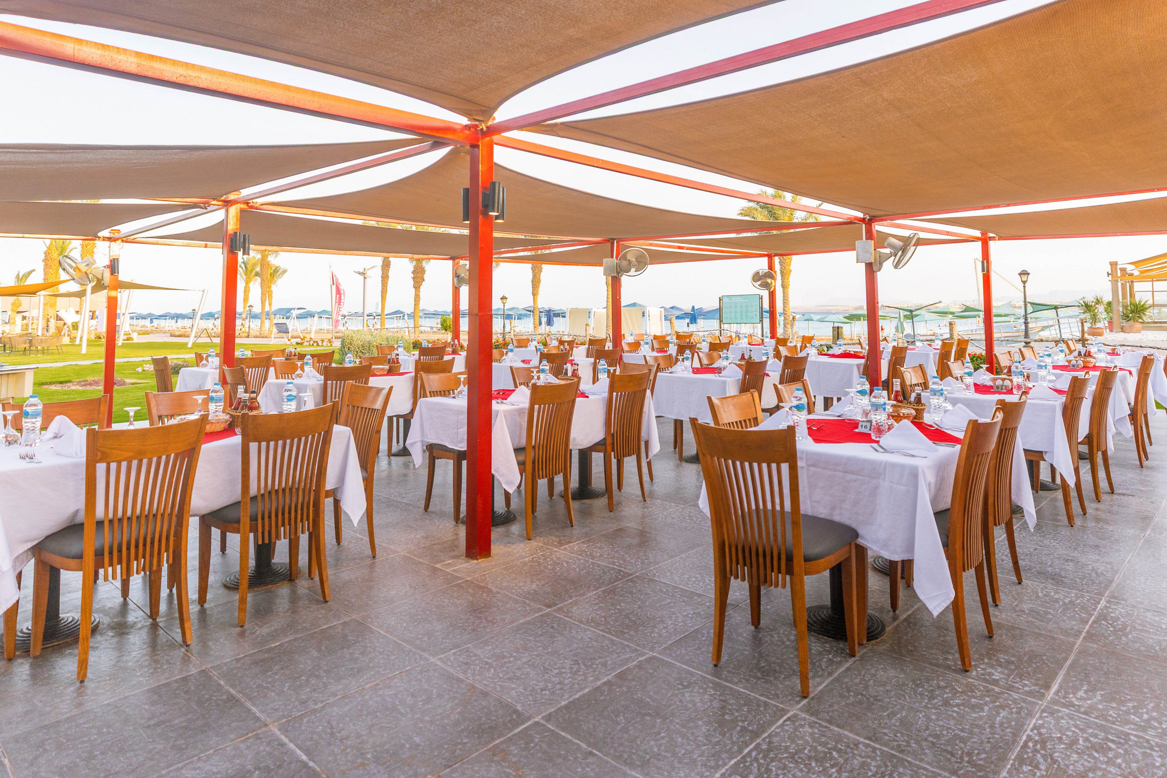 An open-air restaurant features neatly arranged tables covered with white cloths and red napkins. The setting is bright and inviting, with wooden chairs and a shaded canopy overhead. The restaurant overlooks a scenic seaside area, creating a relaxed and elegant dining atmosphere. No visible text or numbers are present in the image.