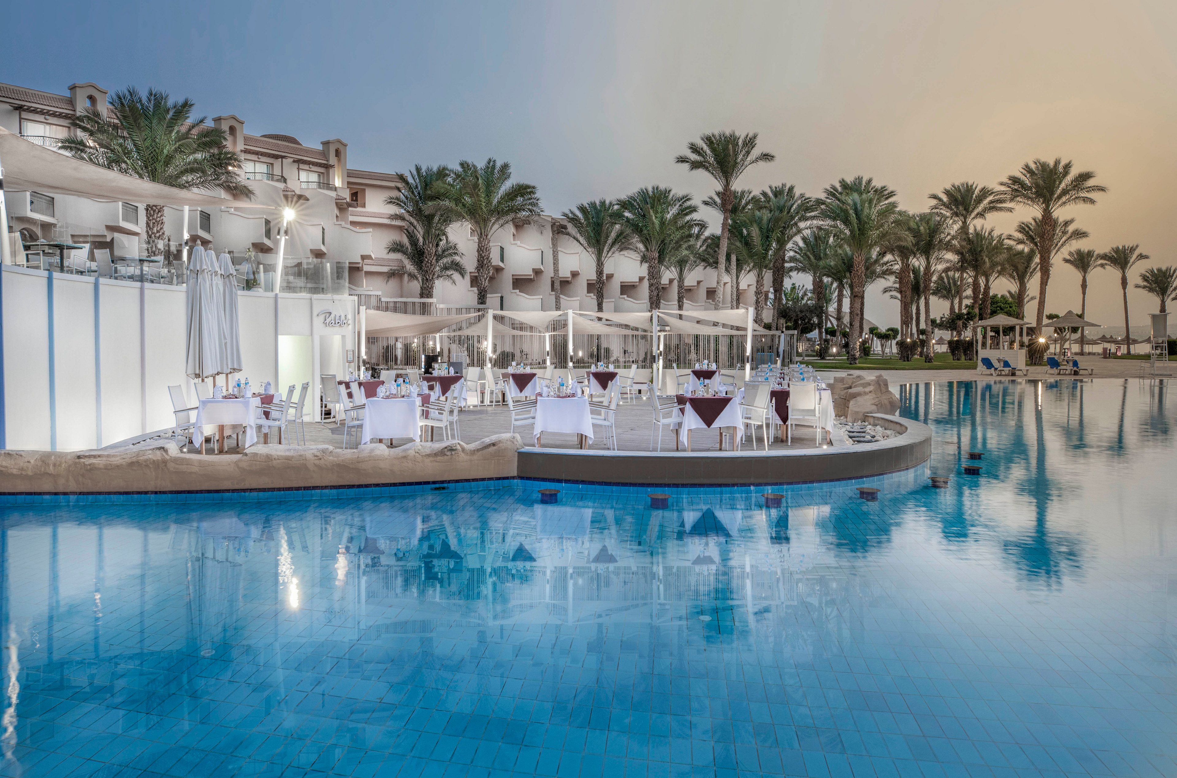 A serene outdoor dining area is set up beside a large swimming pool at a luxury resort. Tables with white linens and chairs are arranged under canopies, surrounded by palm trees and modern architecture. The tranquil blue water reflects the evening sky, creating a relaxing and elegant atmosphere. No visible text or numbers are present in the image.