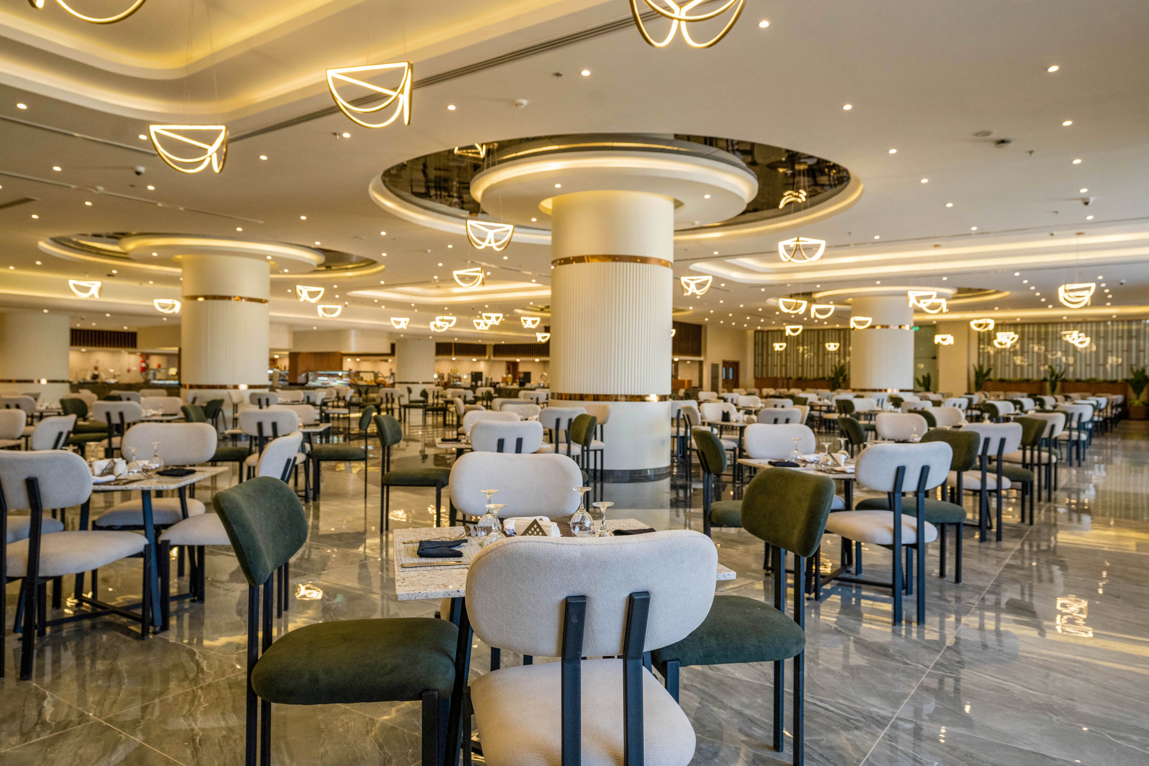 Large contemporary restaurant interior with rows of neatly arranged tables and chairs. The space features polished marble floors, round columns, and decorative circular ceiling lights. Place settings with glassware and cutlery are prepared on each table, suggesting readiness for guests. Warm ambient lighting and neutral tones create an elegant, upscale dining atmosphere.