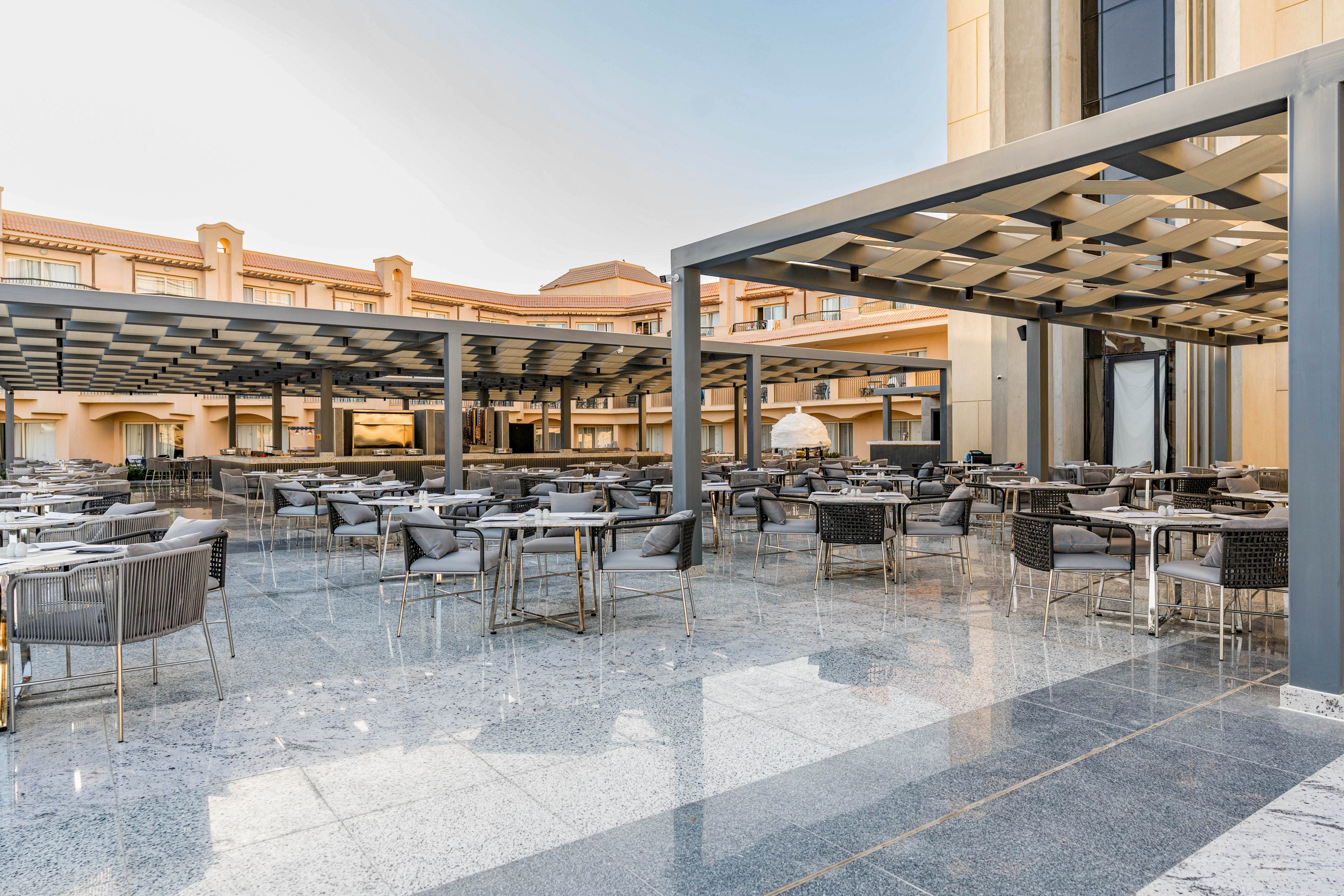 Spacious outdoor dining area with numerous tables and chairs is set on a polished stone terrace beside a large building. Metal pergolas provide partial shade over the seating, creating a structured, contemporary look. The surrounding architecture features warm beige tones and arched details, suggesting a hotel or resort setting. The scene is empty of people, emphasizing the clean, orderly arrangement and bright daylight atmosphere.