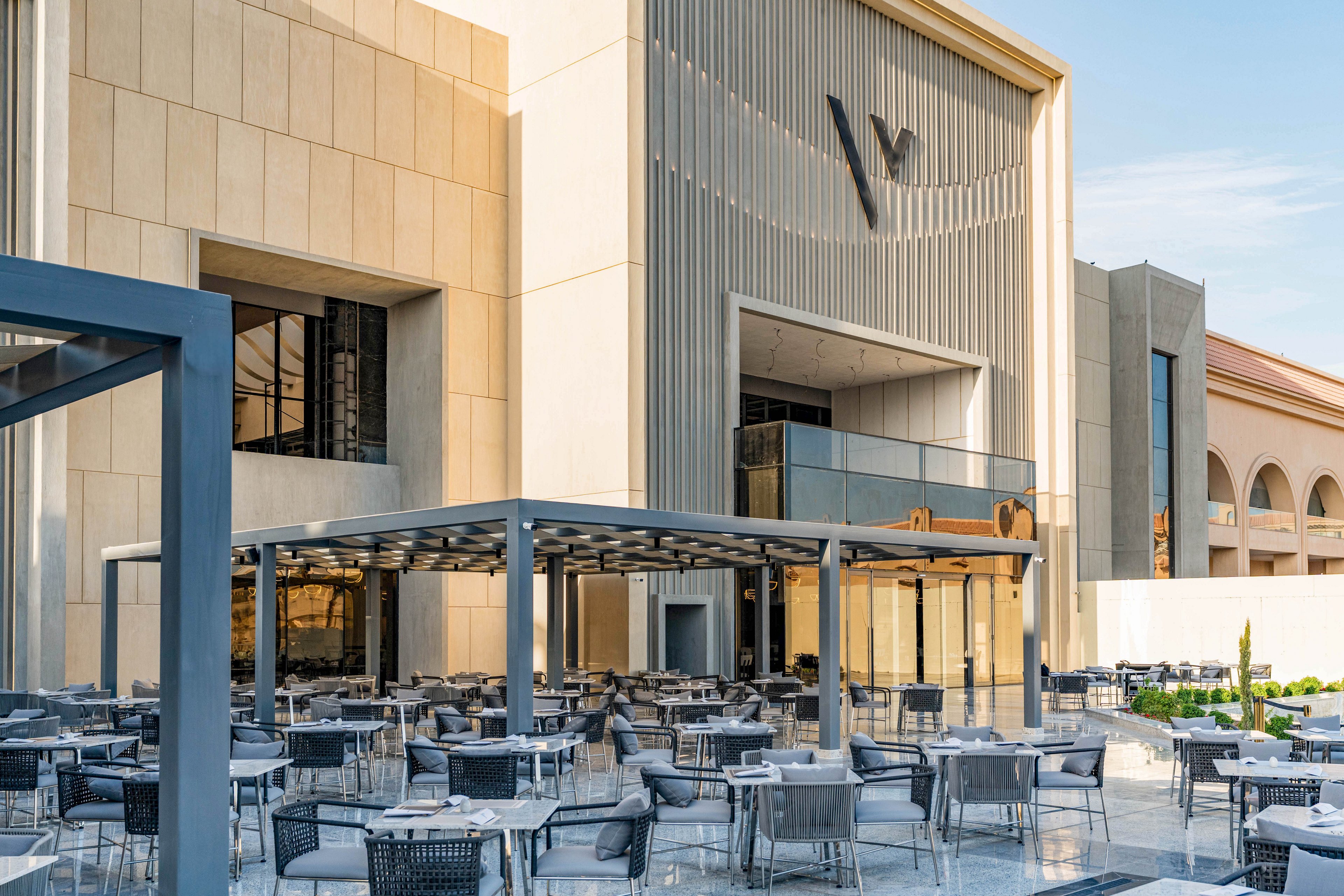 A contemporary outdoor dining area with numerous tables and chairs is set in front of a large, modern building. The architecture features clean lines, tall vertical panels, and a prominent stylized "W" on the facade. A metal pergola structure provides partial shade over the terrace, while glass doors lead into the interior space. Warm sunlight and clear skies create an inviting atmosphere for shoppers and diners.