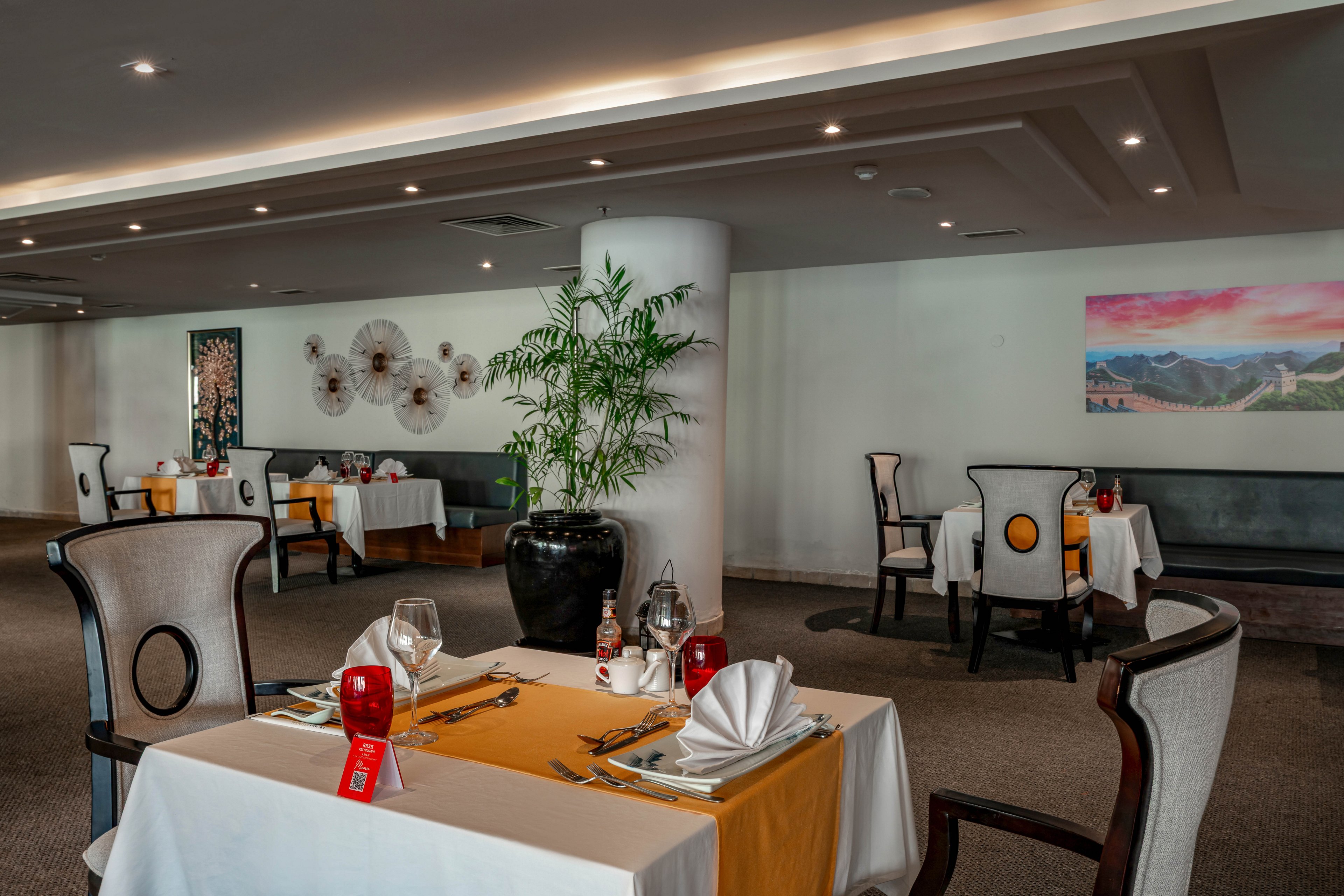 A contemporary restaurant dining area features neatly set tables with white linens, folded napkins, and glassware. The setting is spacious and well-lit, with decorative wall art and a large potted plant adding to the ambiance. The color palette is neutral with accents of yellow and red, creating a welcoming and refined atmosphere. No people are present, emphasizing the calm and orderly environment. Visible table numbers and utensils highlight attention to detail.