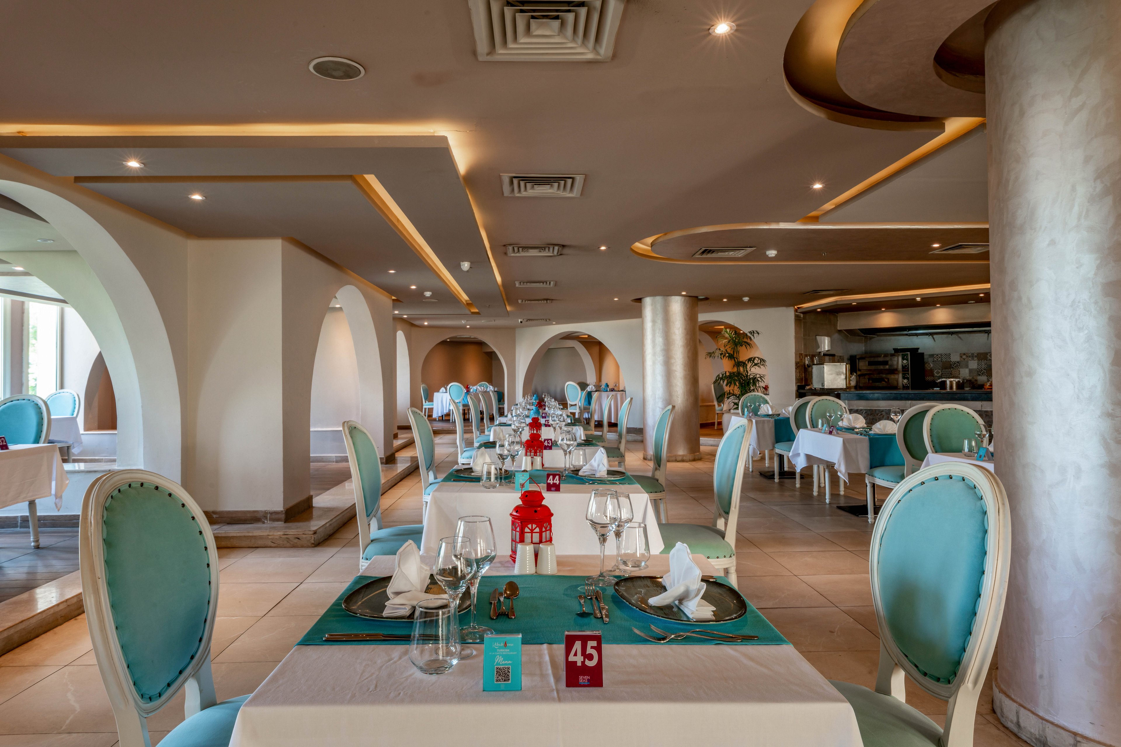 A spacious, upscale restaurant dining area features turquoise upholstered chairs and neatly set tables with white linens and glassware. The setting is bright and contemporary, with arched architectural details and soft ambient lighting. Table number '45' is clearly visible on one of the tables, adding a numeric reference. The overall mood is refined and inviting, suitable for fine dining experiences.