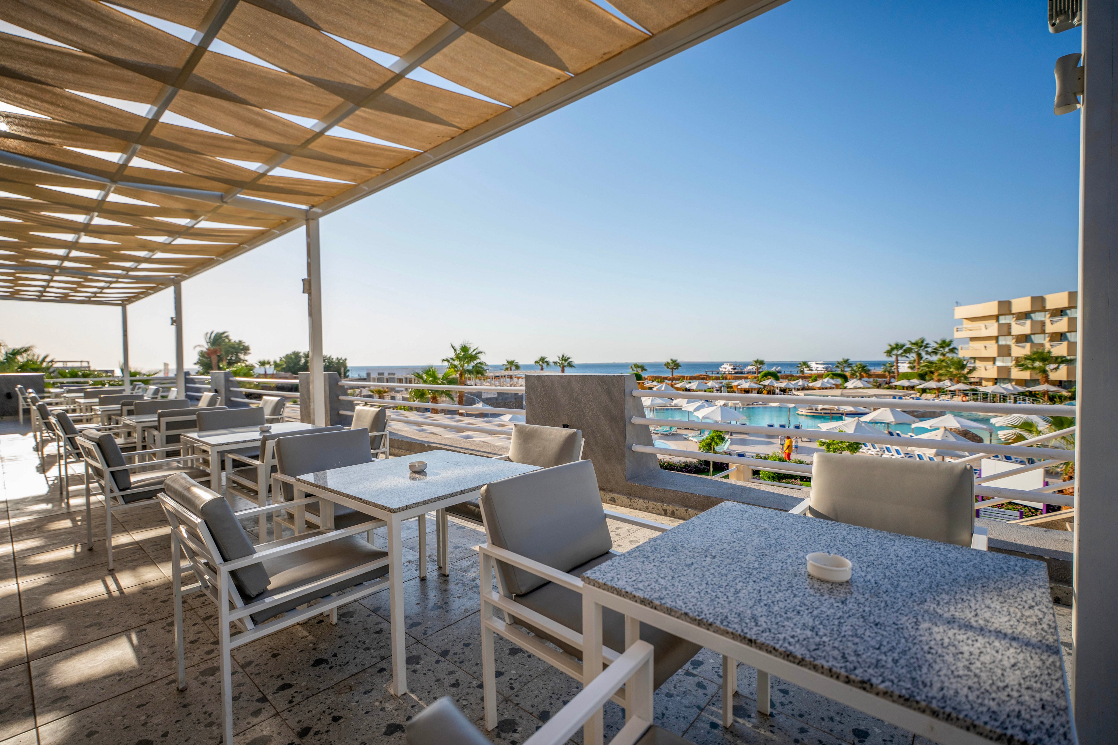 A spacious outdoor terrace features contemporary tables and chairs under a wooden pergola. The setting overlooks a vibrant resort pool area with sun loungers, umbrellas, and palm trees, all set against a clear blue sky. The scene is bright and inviting, evoking a relaxed vacation atmosphere with clean lines and neutral tones. No visible text or numbers are present in the image.