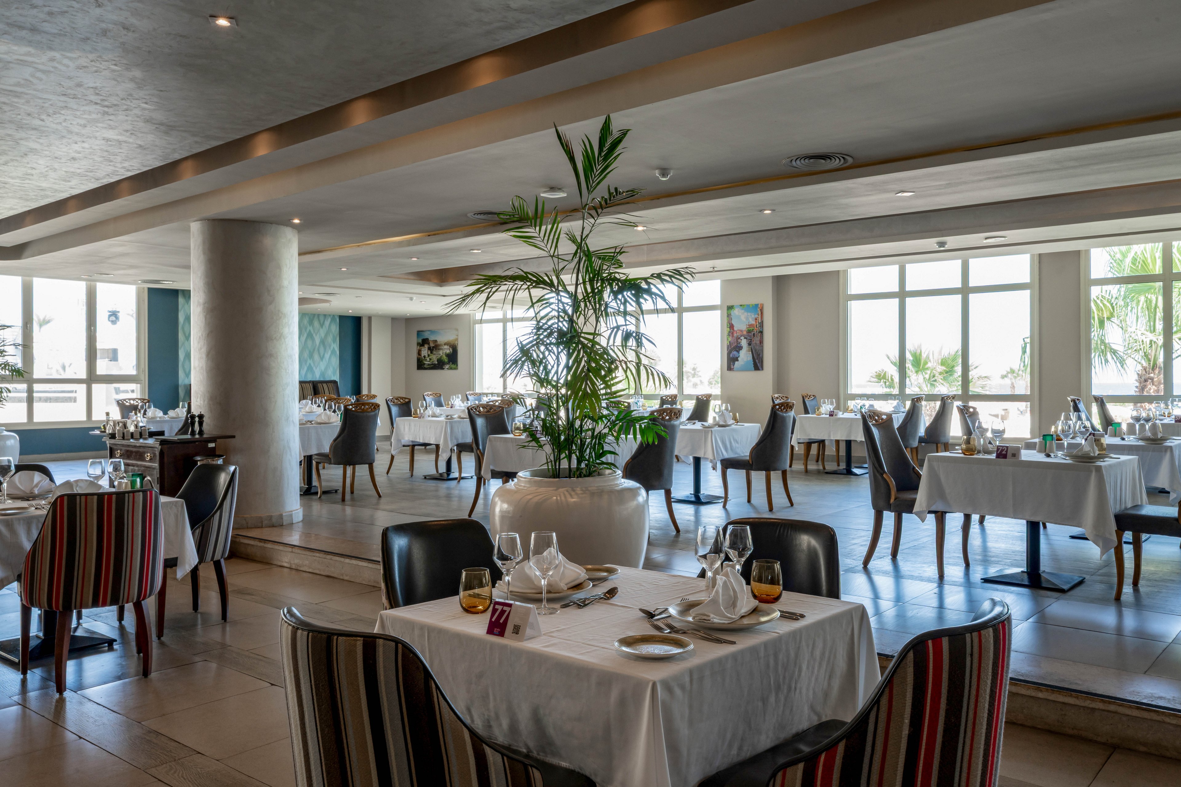A spacious, contemporary restaurant dining area features neatly arranged tables with white tablecloths and formal place settings. Large windows allow natural light to fill the room, highlighting a central decorative plant and striped upholstered chairs. The atmosphere is calm and inviting, with a neutral color palette and a touch of greenery. No visible text or numbers are present in the image.