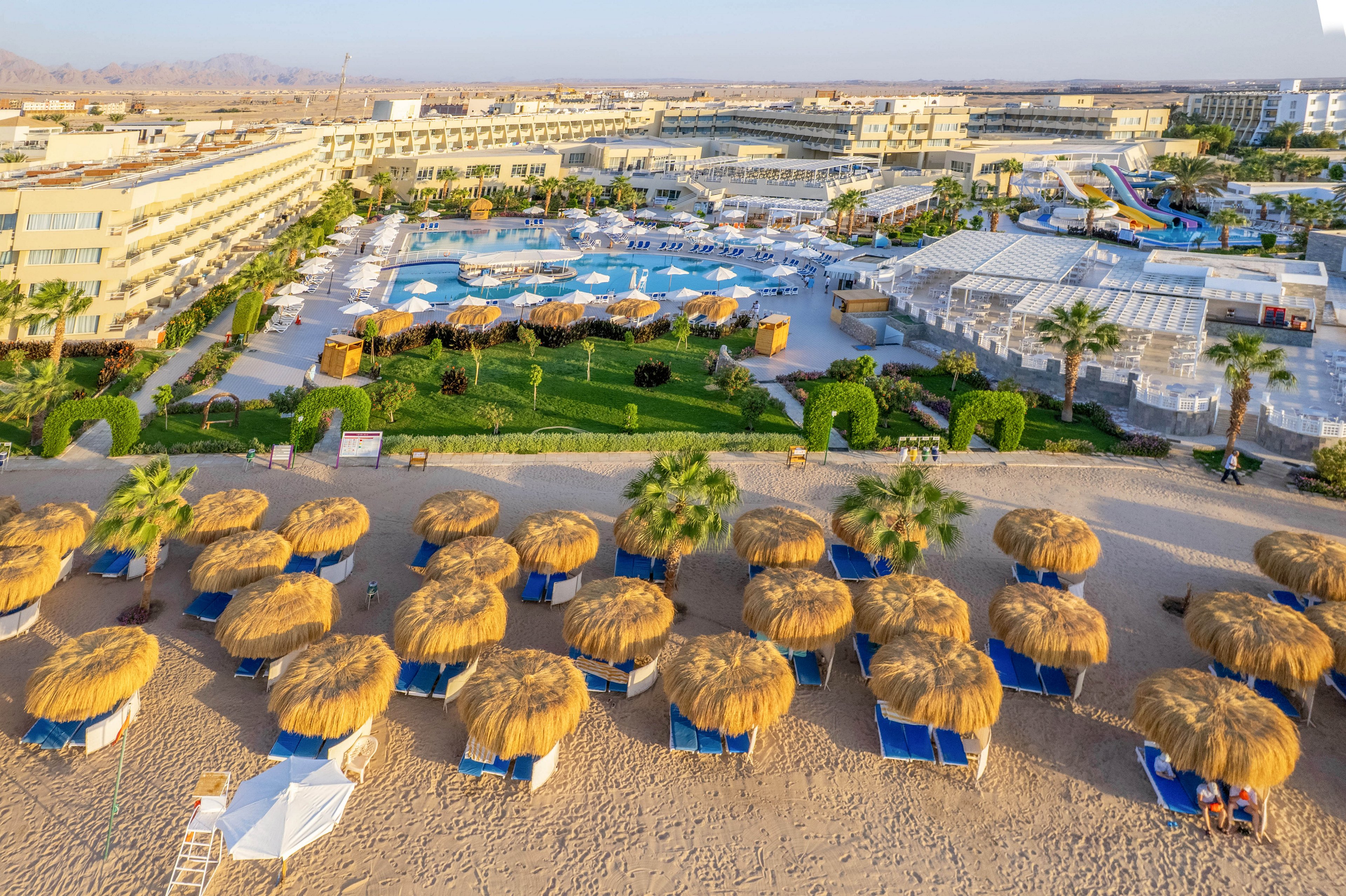 This image shows a large beachfront resort with rows of straw umbrellas and lounge chairs on the sandy shore. The resort features multiple swimming pools, landscaped gardens, and water slides, with several buildings in the background. The setting is sunny and vibrant, evoking a relaxed vacation atmosphere. No visible text or numbers are present in the image.