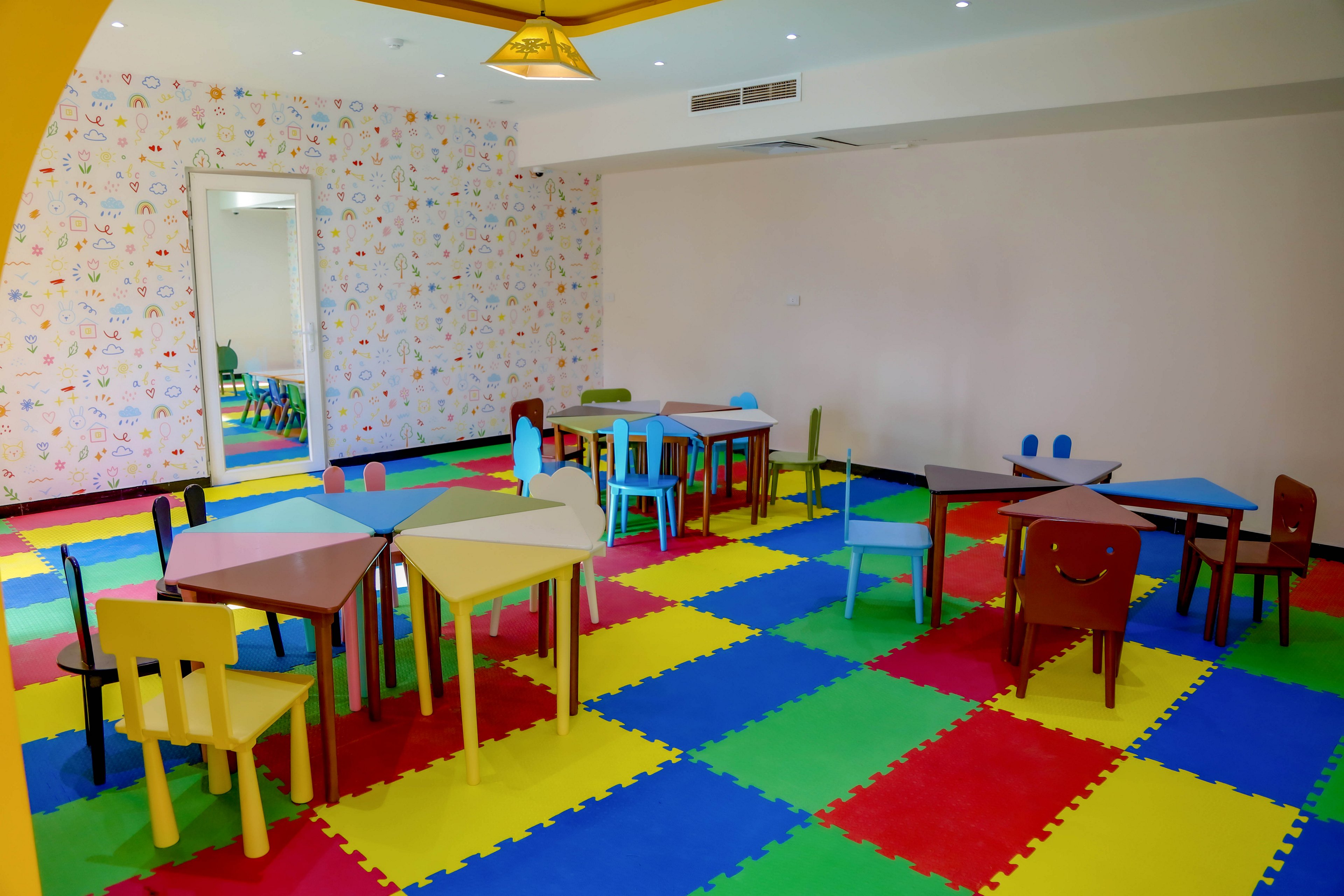 A brightly lit classroom features colorful interlocking foam floor tiles and small, multi-colored tables and chairs arranged throughout the room. The walls are decorated with playful patterns, creating a cheerful and inviting atmosphere. No people are present, and the space appears ready for young children. The overall mood is vibrant and playful, ideal for early childhood learning or daycare settings.
