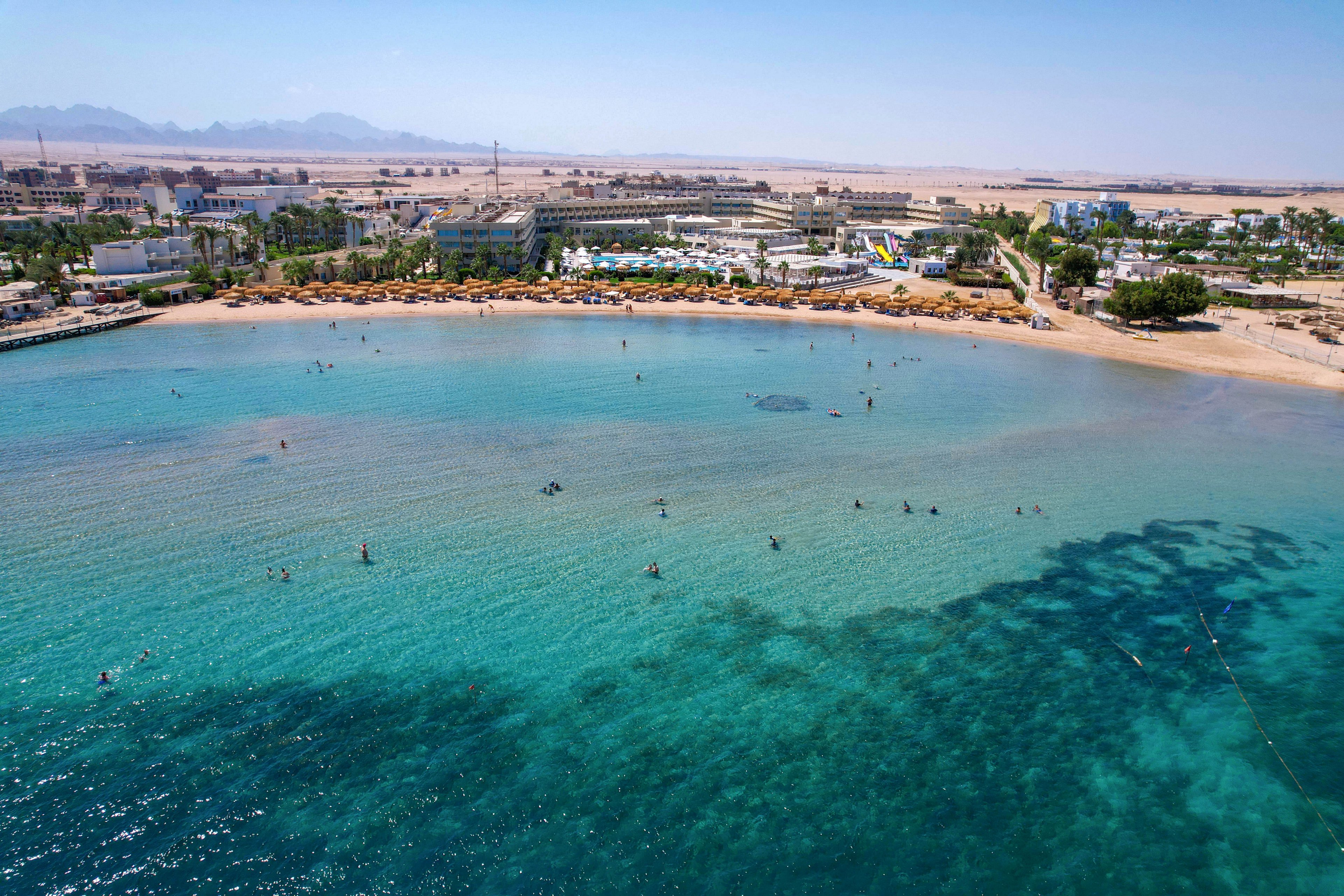 This image captures an aerial perspective of a coastal resort area with a sandy beach and turquoise waters. Numerous people are visible swimming and paddleboarding in the calm bay. The shoreline is lined with umbrellas and sun loungers, while the background features hotels, palm trees, and desert landscape. The overall mood is relaxed and summery, with clear skies and vibrant blue-green water.