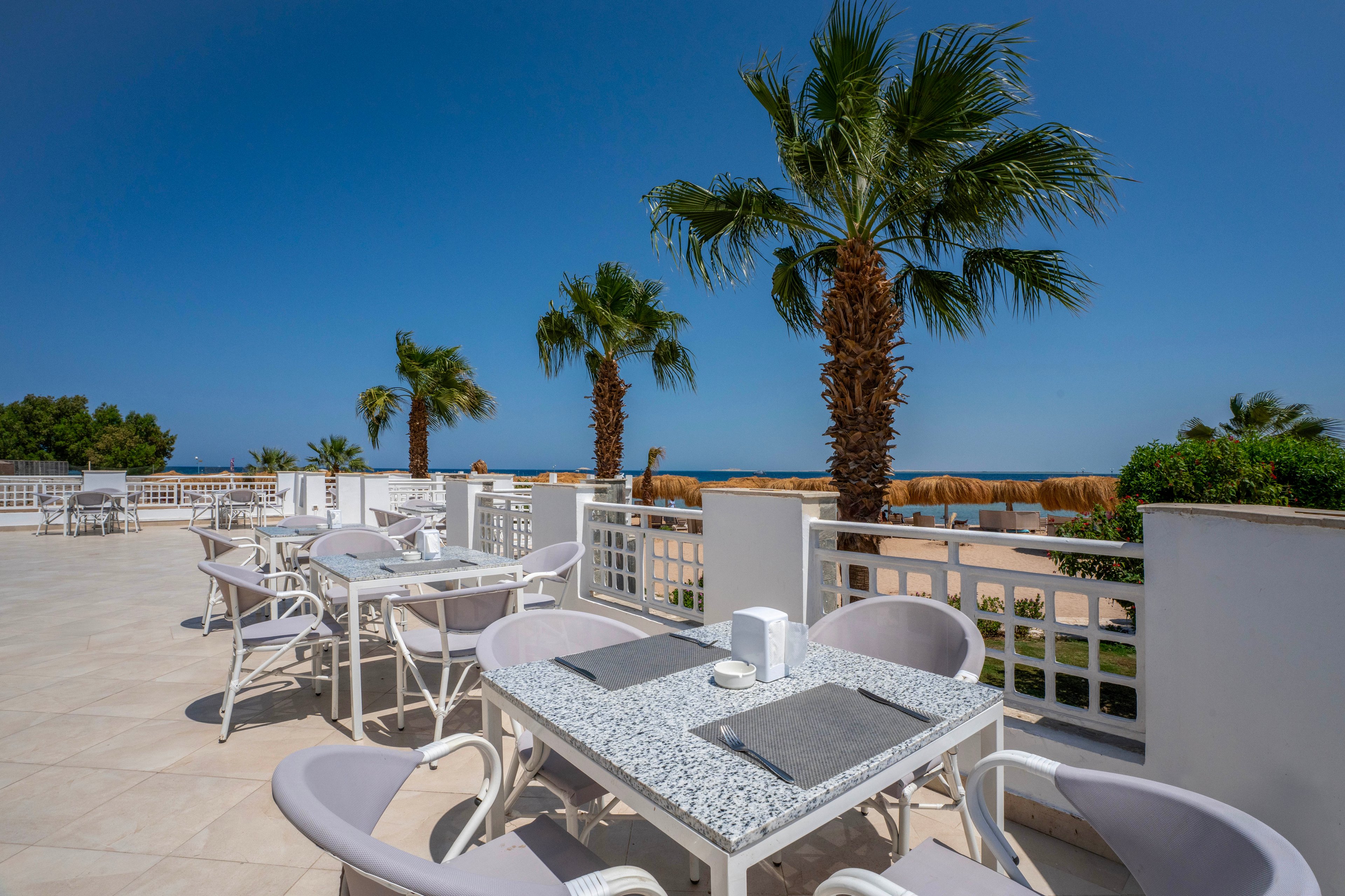 A spacious outdoor terrace features several white tables and chairs arranged neatly under a clear blue sky. Tall palm trees line the edge of the terrace, offering a tropical atmosphere with a scenic view of the ocean in the background. The setting is bright and inviting, with a modern, minimalist style and a calm, relaxing mood. No people are present, emphasizing the tranquil environment.