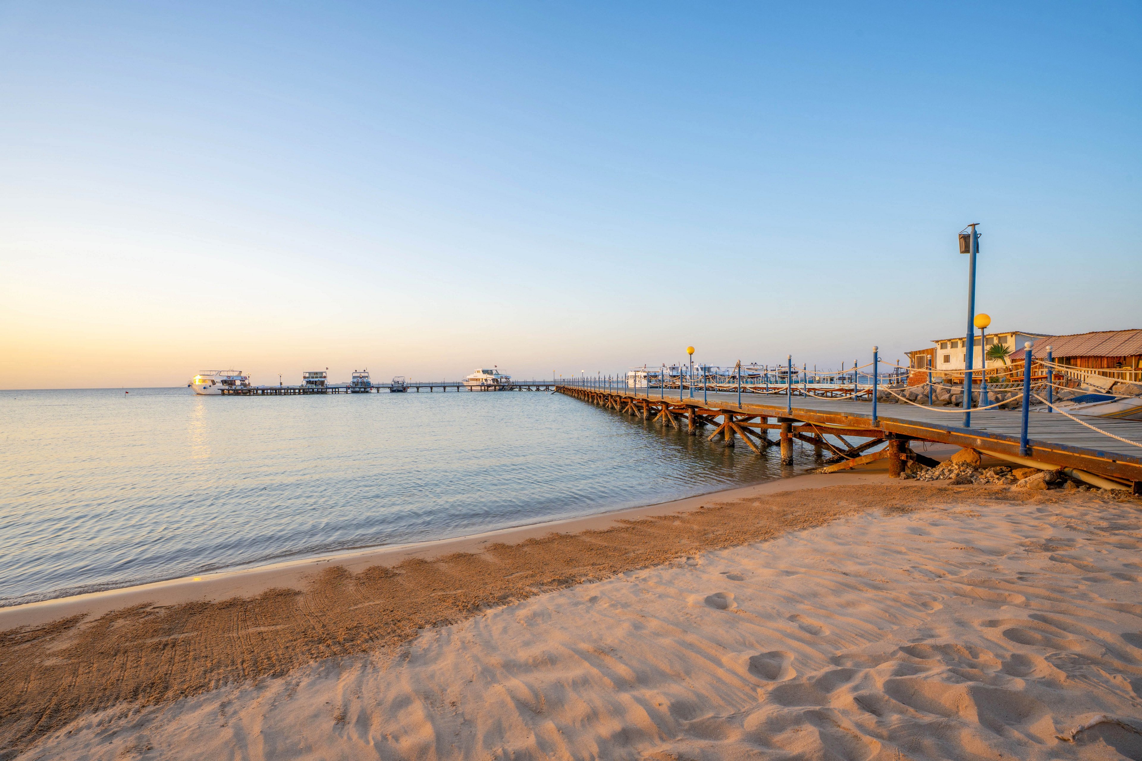 A long wooden pier extends over tranquil water, leading out from a sandy beach under a clear blue sky. The scene is bathed in warm early morning light, creating a peaceful and inviting atmosphere. Several small structures are visible at the end of the pier, with gentle waves lapping at the shore. The setting suggests a serene coastal location, ideal for relaxation or leisure activities.