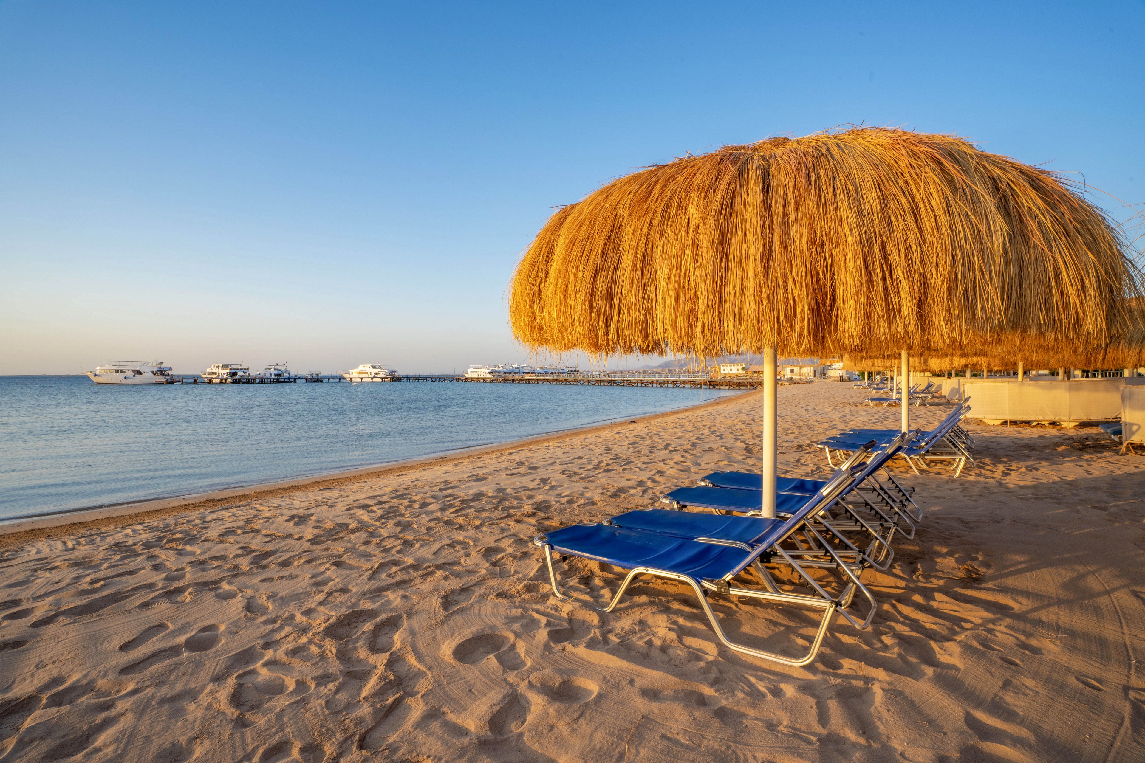 A tranquil sandy beach features rows of blue lounge chairs under large straw umbrellas. The calm sea stretches out to the horizon, with several boats anchored near a distant pier. The scene is bathed in warm, golden sunlight, evoking a relaxed and peaceful vacation atmosphere. No visible people or text are present in the image.