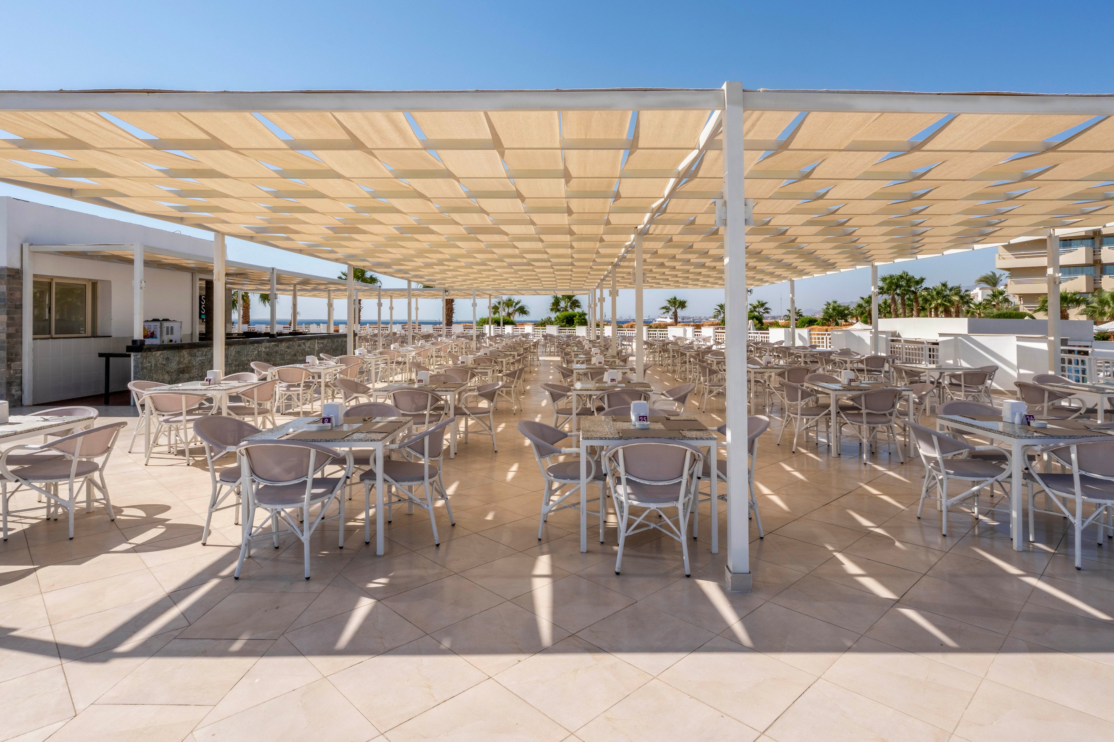 A spacious outdoor dining terrace features numerous empty tables and chairs arranged in neat rows under a large, modern pergola. The setting is bright and sunlit, with a tiled floor and a clear blue sky overhead. The area appears ready for guests, with palm trees and buildings visible in the background, creating a relaxed and inviting atmosphere.