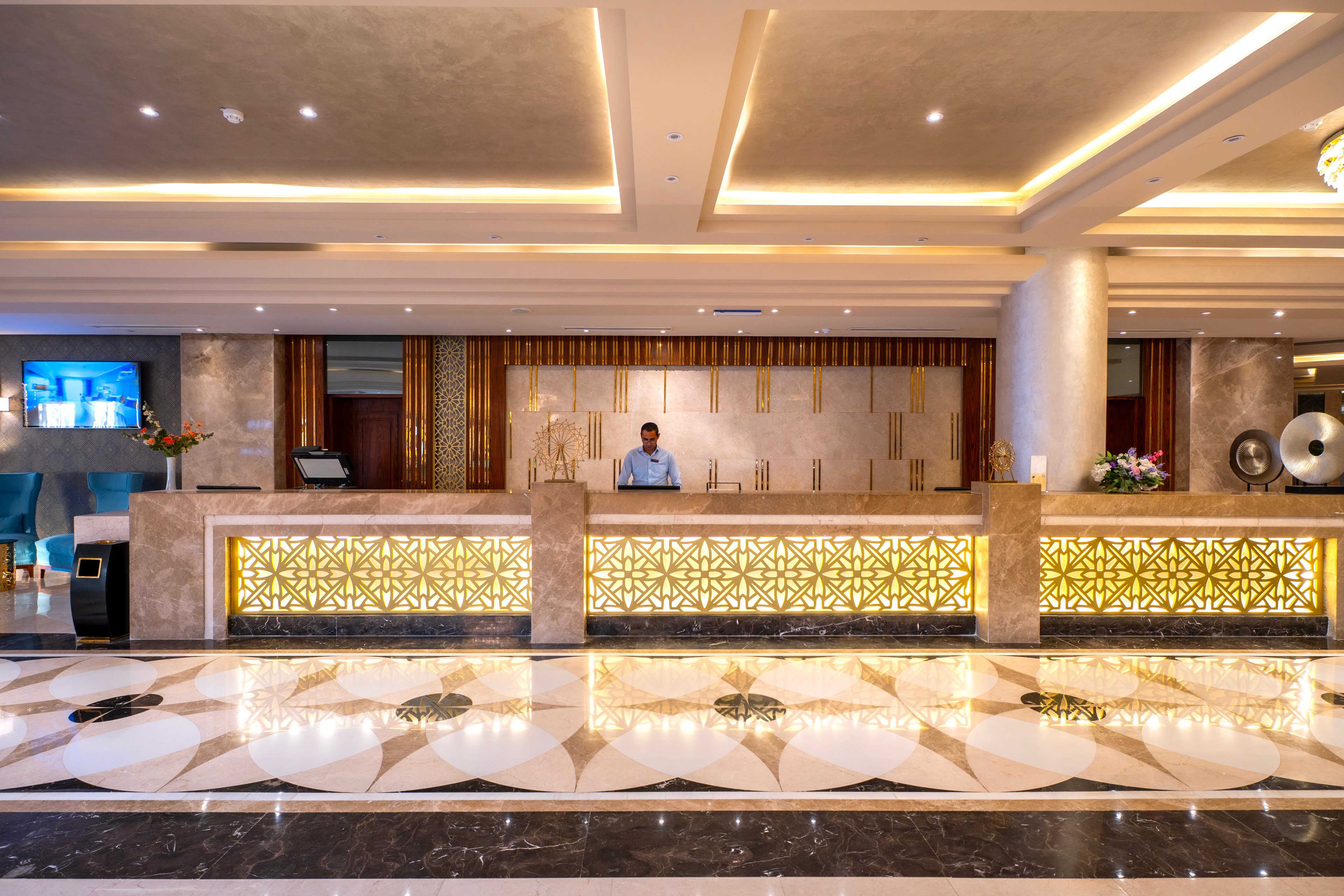 A spacious hotel lobby features a single male staff member standing behind a large, illuminated reception desk. The setting is elegant, with marble floors, decorative lighting, and floral arrangements on the counter. The overall mood is welcoming and luxurious, with warm tones and geometric patterns enhancing the contemporary style. A television screen and computer equipment are visible in the background.