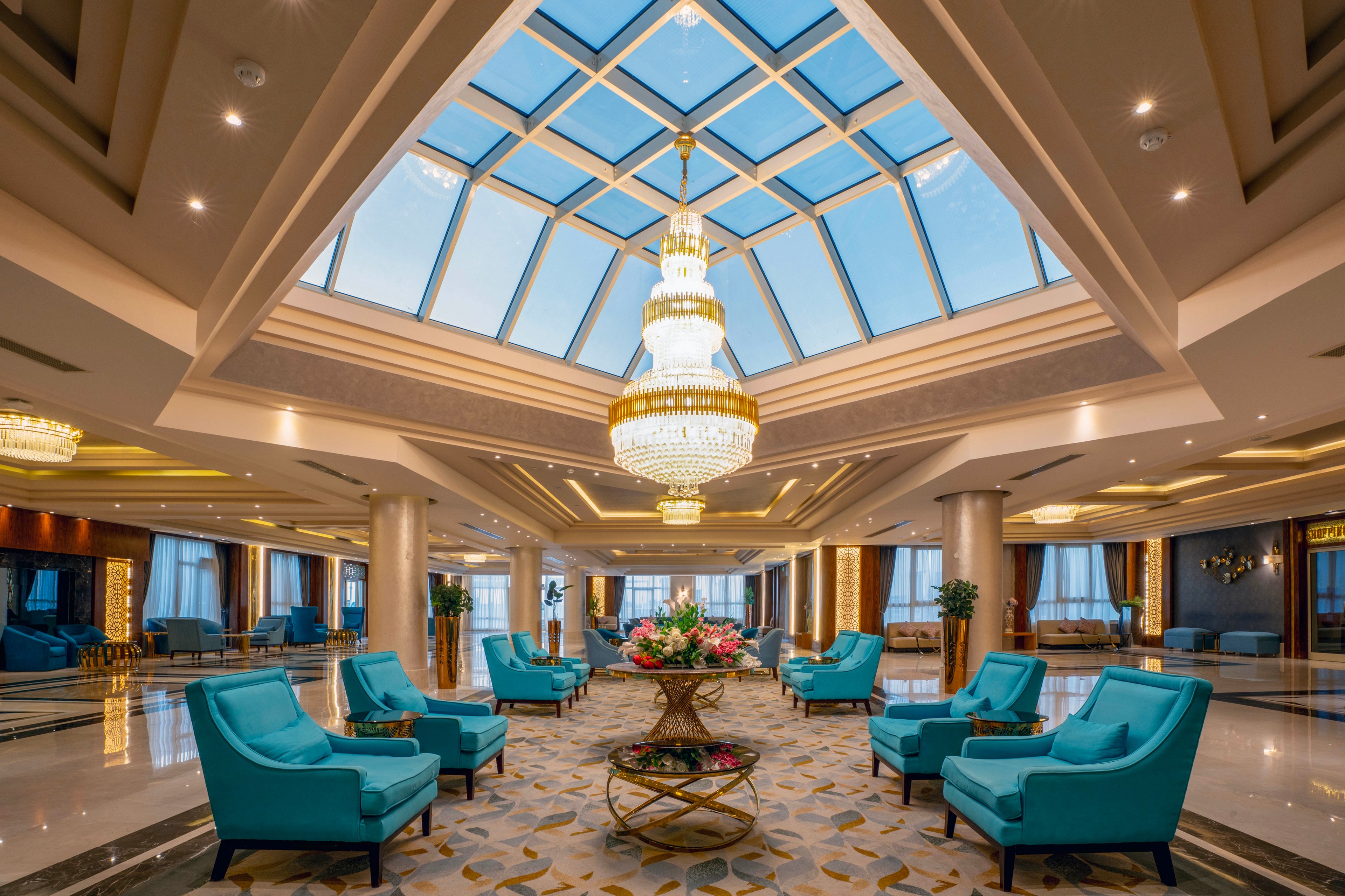 A luxurious hotel lobby features a grand glass skylight ceiling and a large, ornate chandelier as the centerpiece. Plush turquoise armchairs are arranged around a decorative table with a floral arrangement, creating an inviting seating area. The space is illuminated by natural light and elegant interior lighting, with marble floors and gold accents enhancing the sophisticated atmosphere. No visible text or numbers are present in the image.