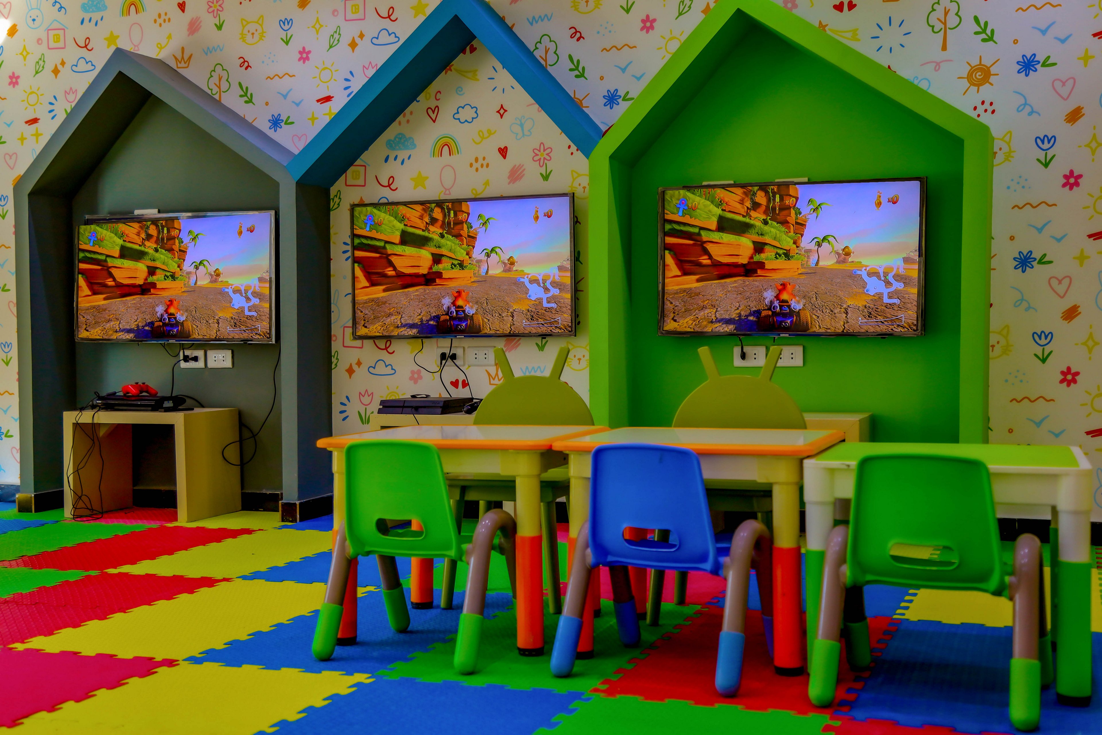 A vibrant children's playroom features three wall-mounted televisions displaying a racing video game. Small colorful tables and chairs are arranged in front of each gaming station, set against a playful patterned wall and foam puzzle flooring. The room is designed for young children, with bright colors and a cheerful atmosphere.