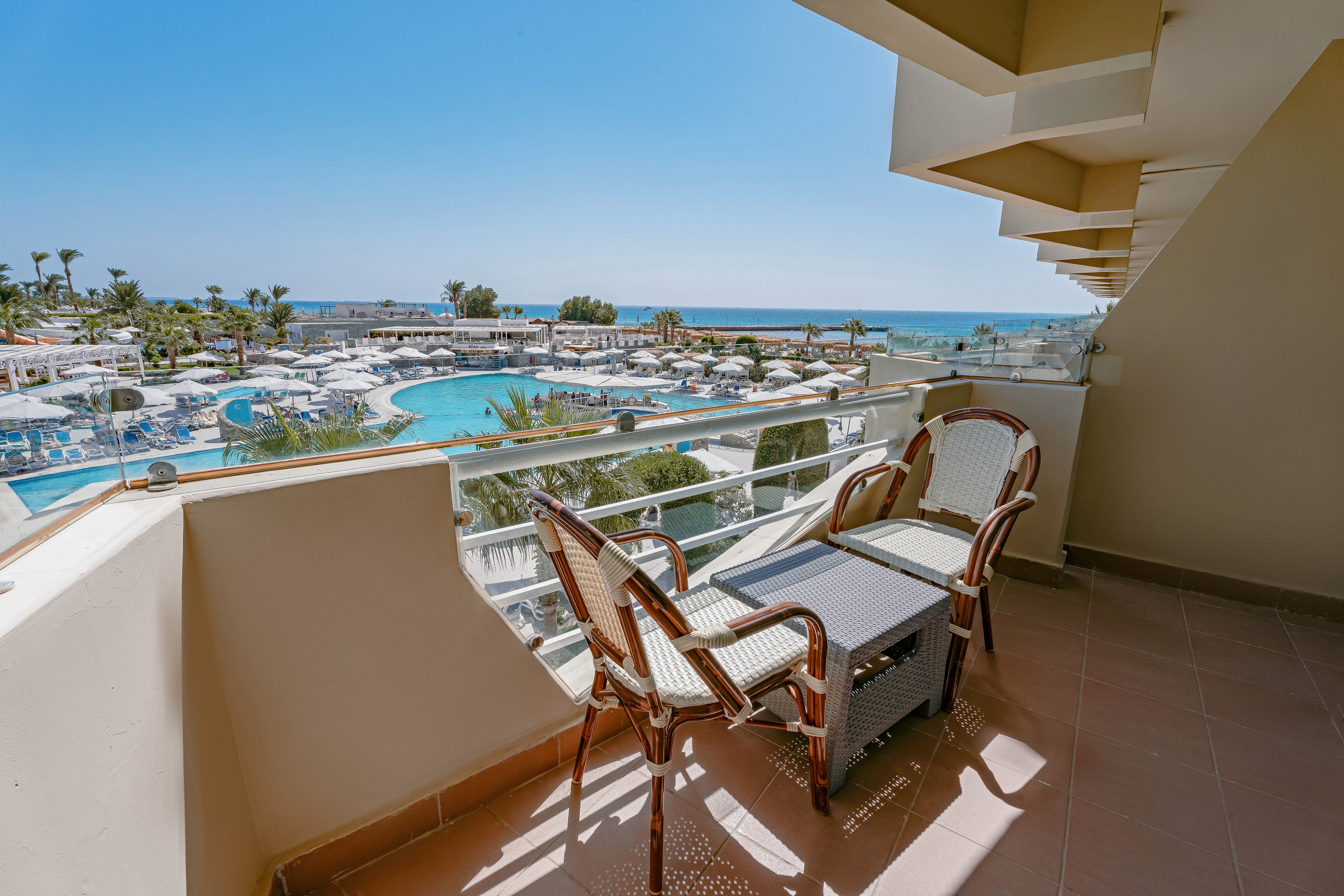 A spacious balcony features a table and four chairs overlooking a large resort pool area and the ocean beyond. The setting is bright and sunny, with clear blue skies and palm trees visible in the distance. The scene conveys a relaxed, vacation atmosphere with modern outdoor furniture and inviting leisure spaces.