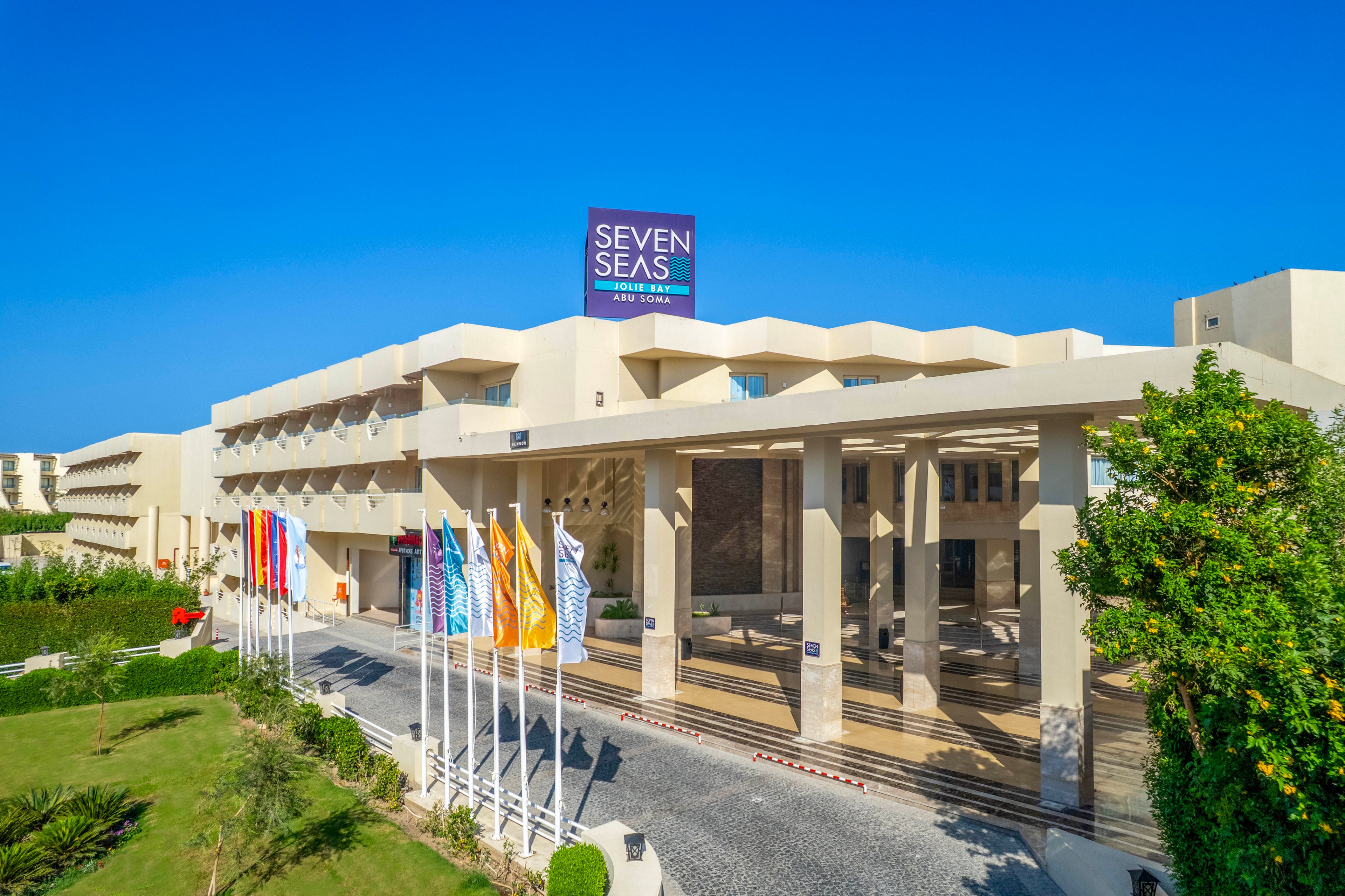 The image shows the exterior of the Seven Seas Resort hotel under a clear blue sky. Multiple international flags are displayed at the entrance, adding a welcoming and global atmosphere. The building features modern architecture with clean lines and beige tones, surrounded by well-maintained greenery. The sign 'SEVEN SEAS RESORT HOTEL' is clearly visible on the rooftop.