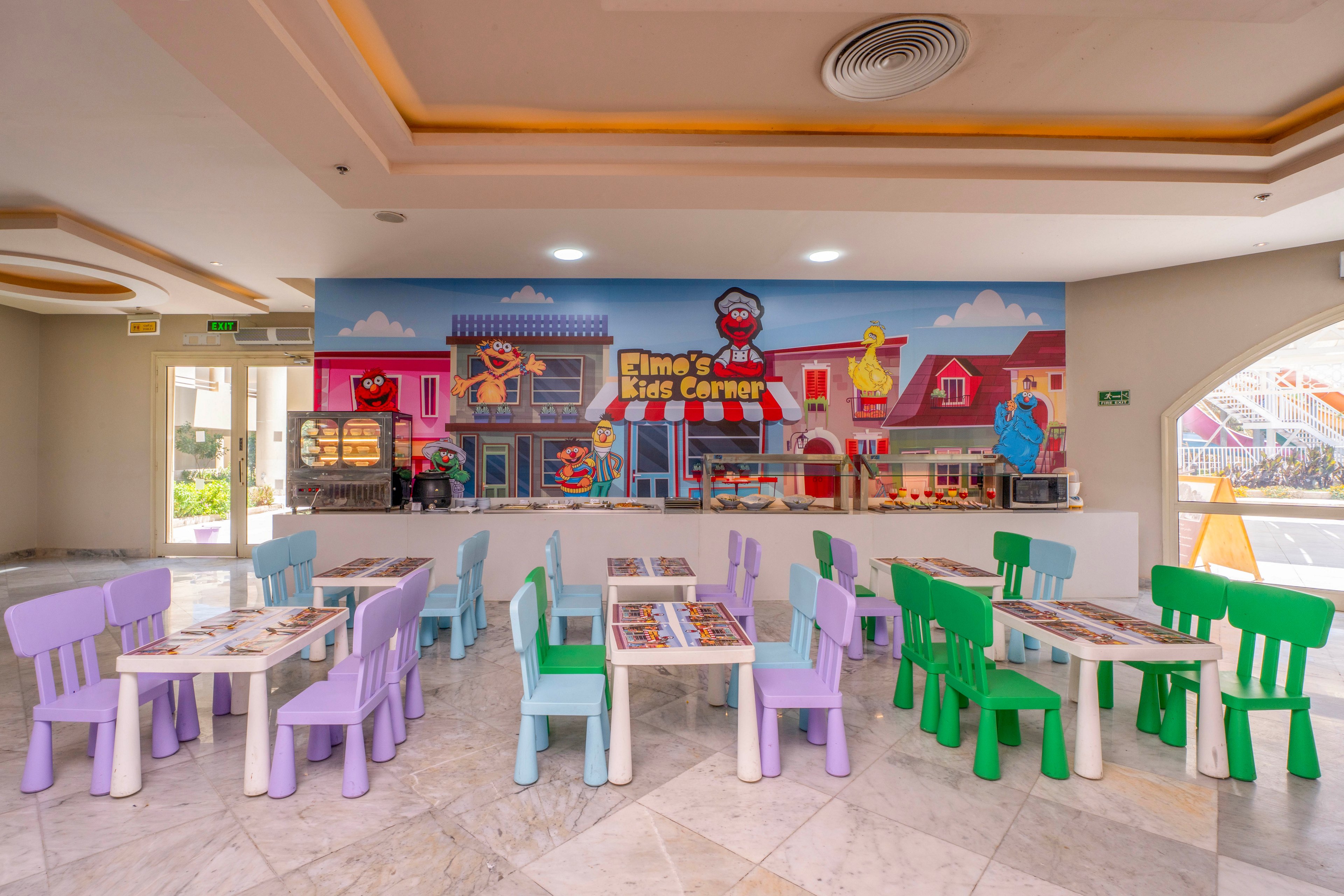A vibrant indoor children's play area features pastel-colored chairs and tables arranged neatly on a marble floor. The back wall displays a large, colorful mural with cartoon characters and the visible text 'Elmo's Kids Corner.' The setting is bright and welcoming, designed for young children, with playful visuals and a cheerful atmosphere.