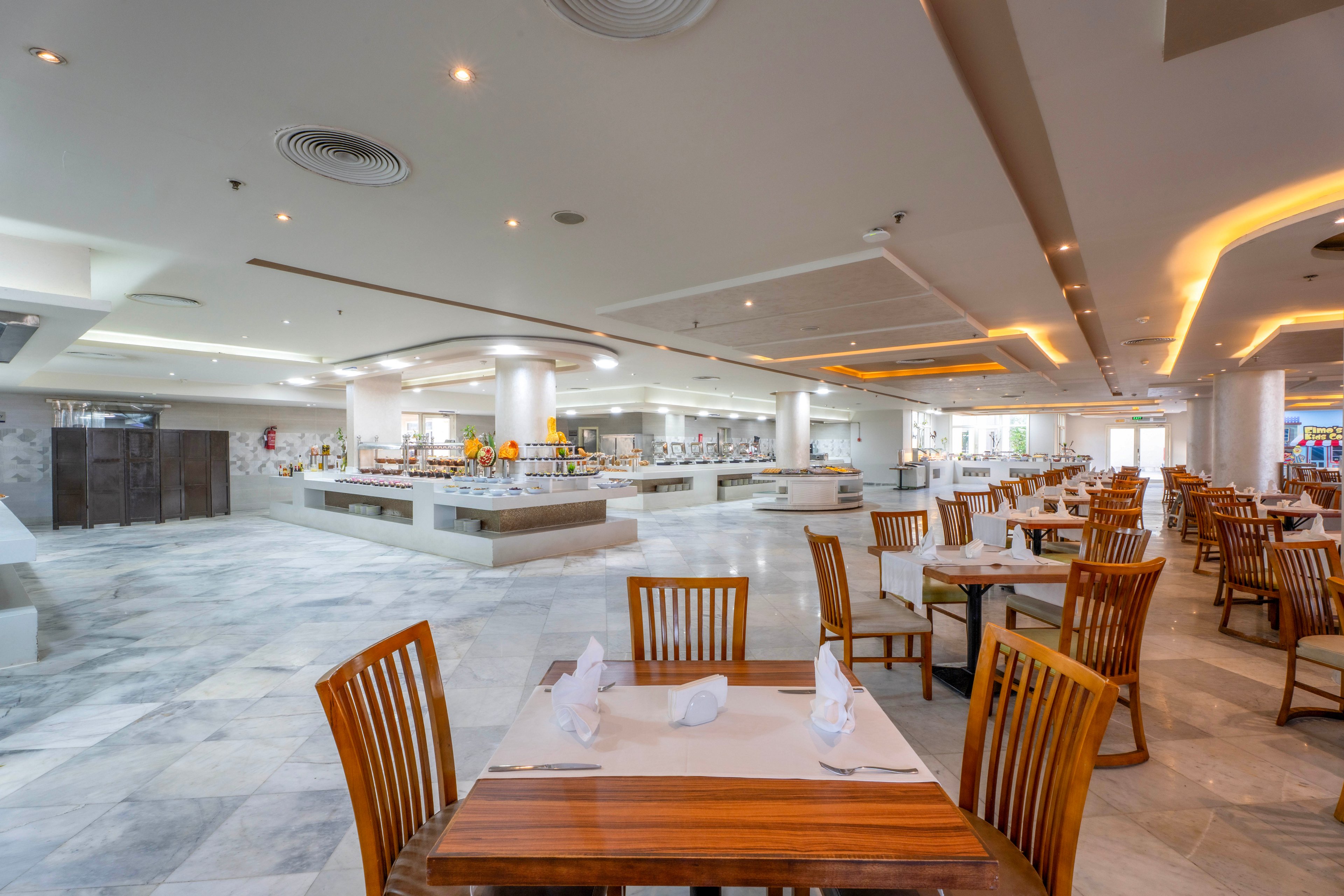 A large, contemporary buffet restaurant features neatly arranged wooden tables and chairs, set with white napkins and tableware. The setting is bright and airy, with a neutral color palette and ample natural light streaming in from large windows. The buffet area is visible in the background, displaying a variety of food stations. The overall mood is clean, inviting, and ready for guests. No visible people or text are present in the image.