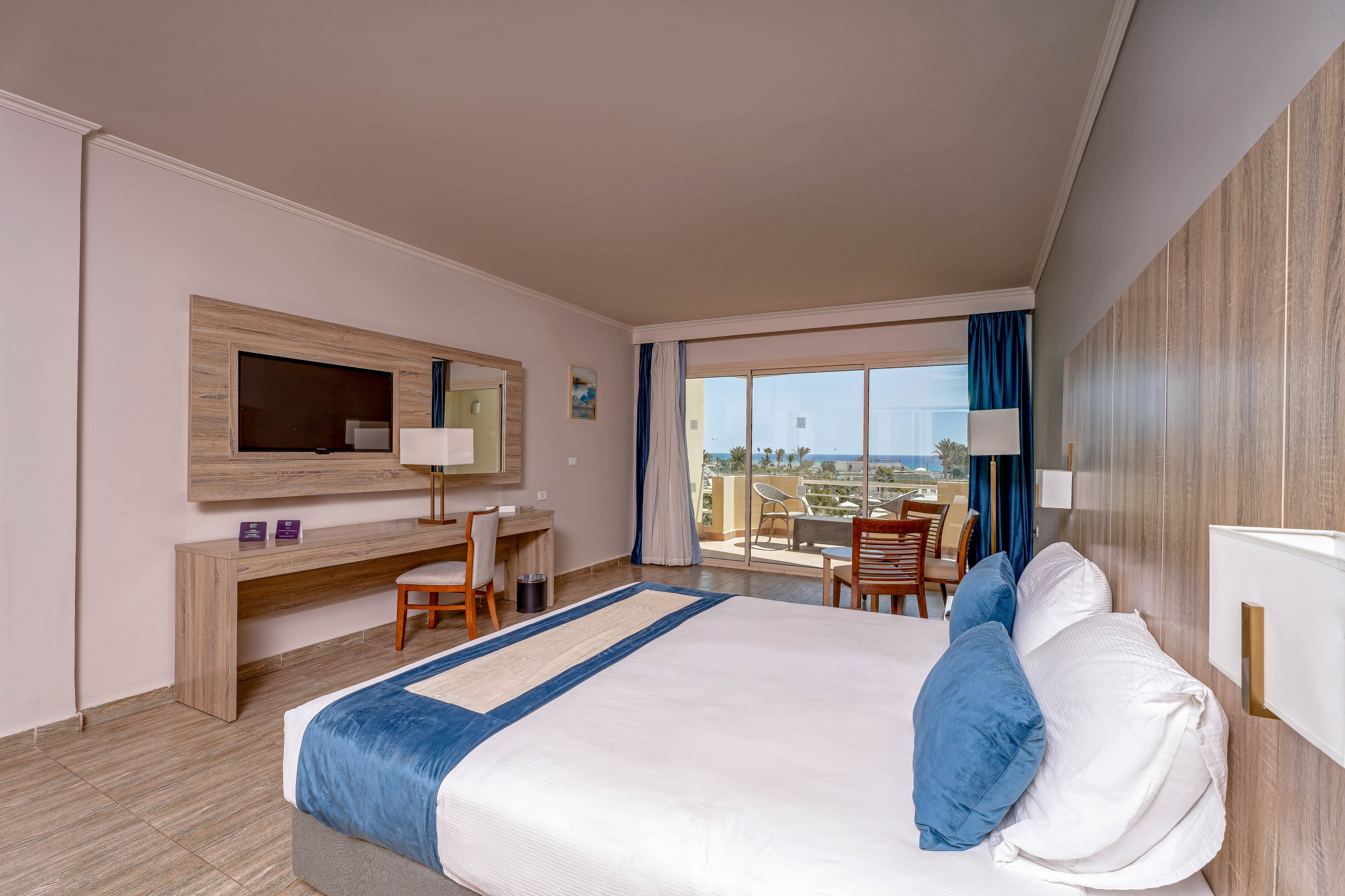 A spacious hotel room features a large bed with blue and white linens, a wooden accent wall, and a flat-screen TV mounted above a desk. The room opens to a balcony with outdoor seating, offering a scenic view of the outdoors. Natural light fills the space, highlighting the contemporary decor and relaxing atmosphere.