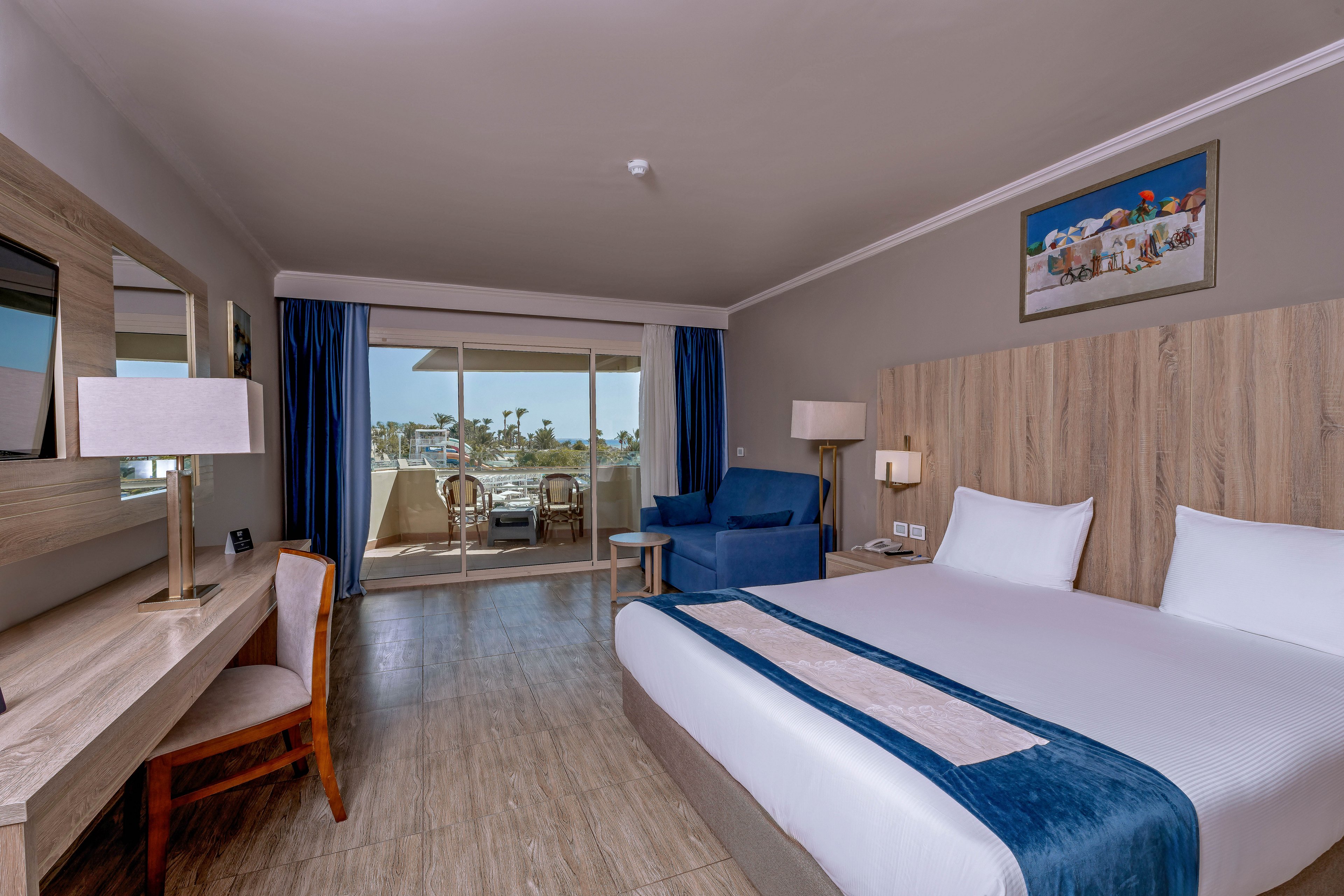 A spacious hotel room features a large bed with white linens and a blue runner, a wooden desk with a lamp, and a comfortable armchair. The room opens onto a balcony with outdoor seating, offering a view of palm trees and a sunny landscape. The decor is contemporary with neutral tones, and a framed artwork hangs above the bed.