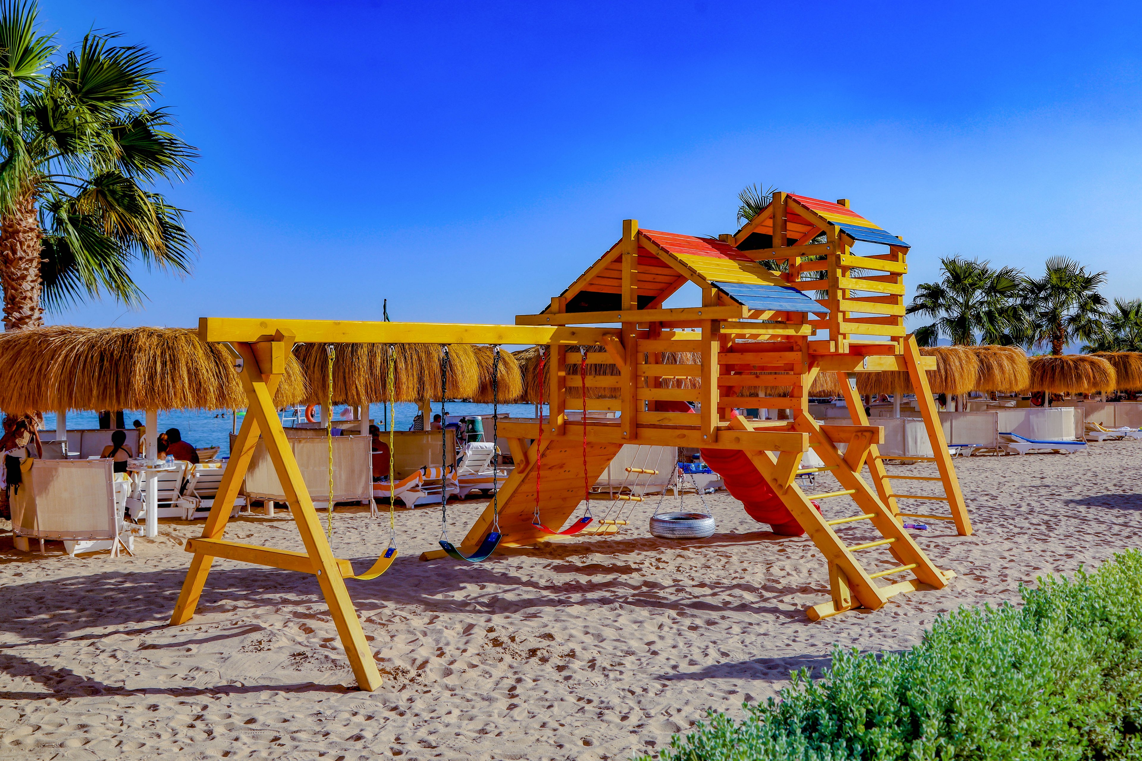 A vibrant wooden playground structure stands on a sandy beach, featuring slides, swings, and climbing areas. The setting is a sunny seaside resort with palm trees and thatched cabanas in the background. The sky is clear and blue, creating a cheerful and inviting atmosphere. No people are visible in the scene, emphasizing the tranquil and family-friendly environment.