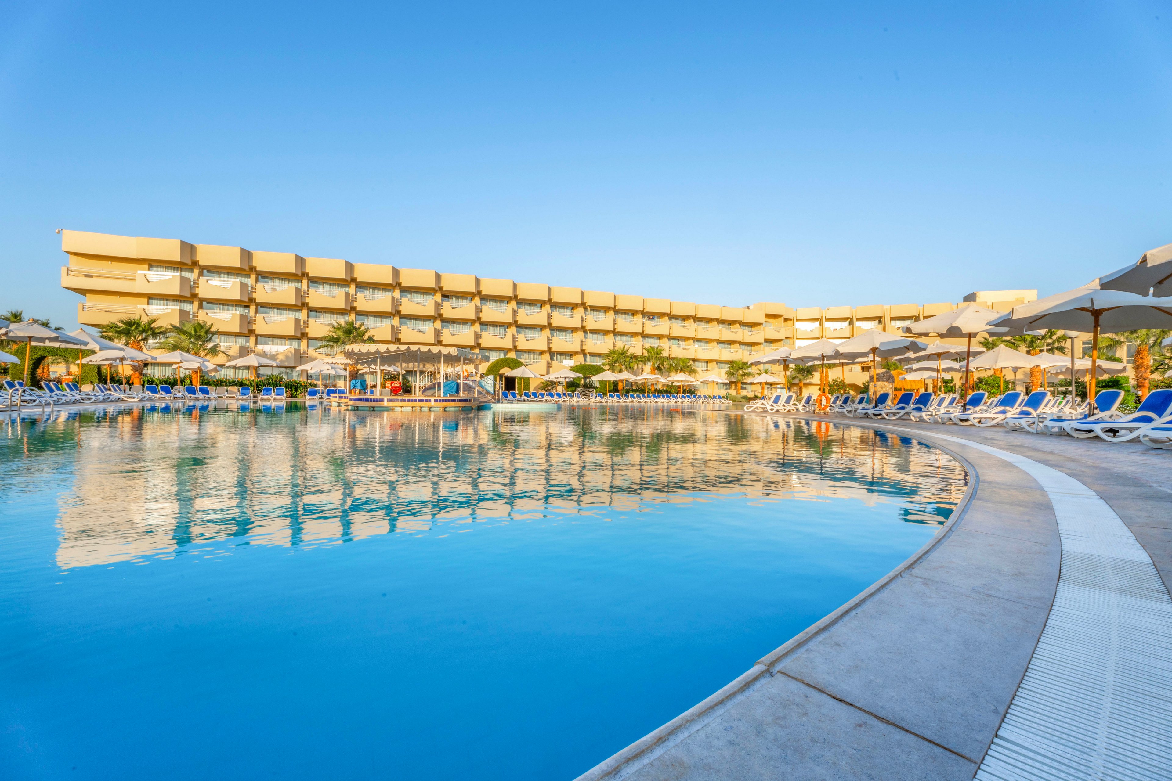 A spacious outdoor swimming pool is featured in front of a contemporary multi-story resort hotel. The scene is set under a clear blue sky, with rows of sun loungers and umbrellas lining the poolside. Palm trees add a tropical touch, and the tranquil water reflects the building and sky, creating a relaxing vacation atmosphere. No visible text or numbers are present in the image.
