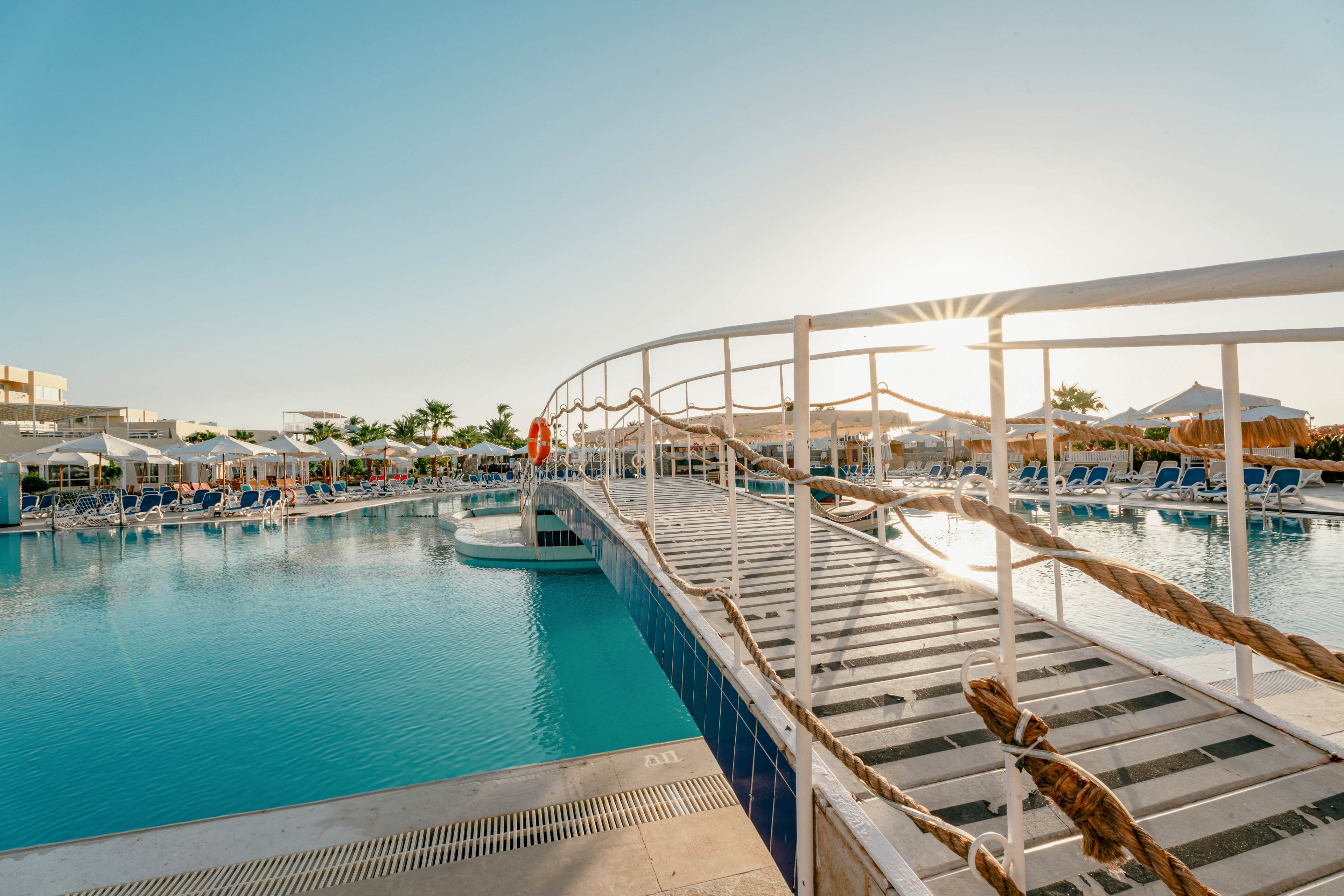 A large outdoor swimming pool is featured at a resort, with a white footbridge crossing over the water. Lounge chairs and umbrellas line the poolside, and a few people are visible enjoying the sunny weather. The scene is set during the day with clear blue skies and palm trees in the background, creating a relaxed vacation atmosphere.