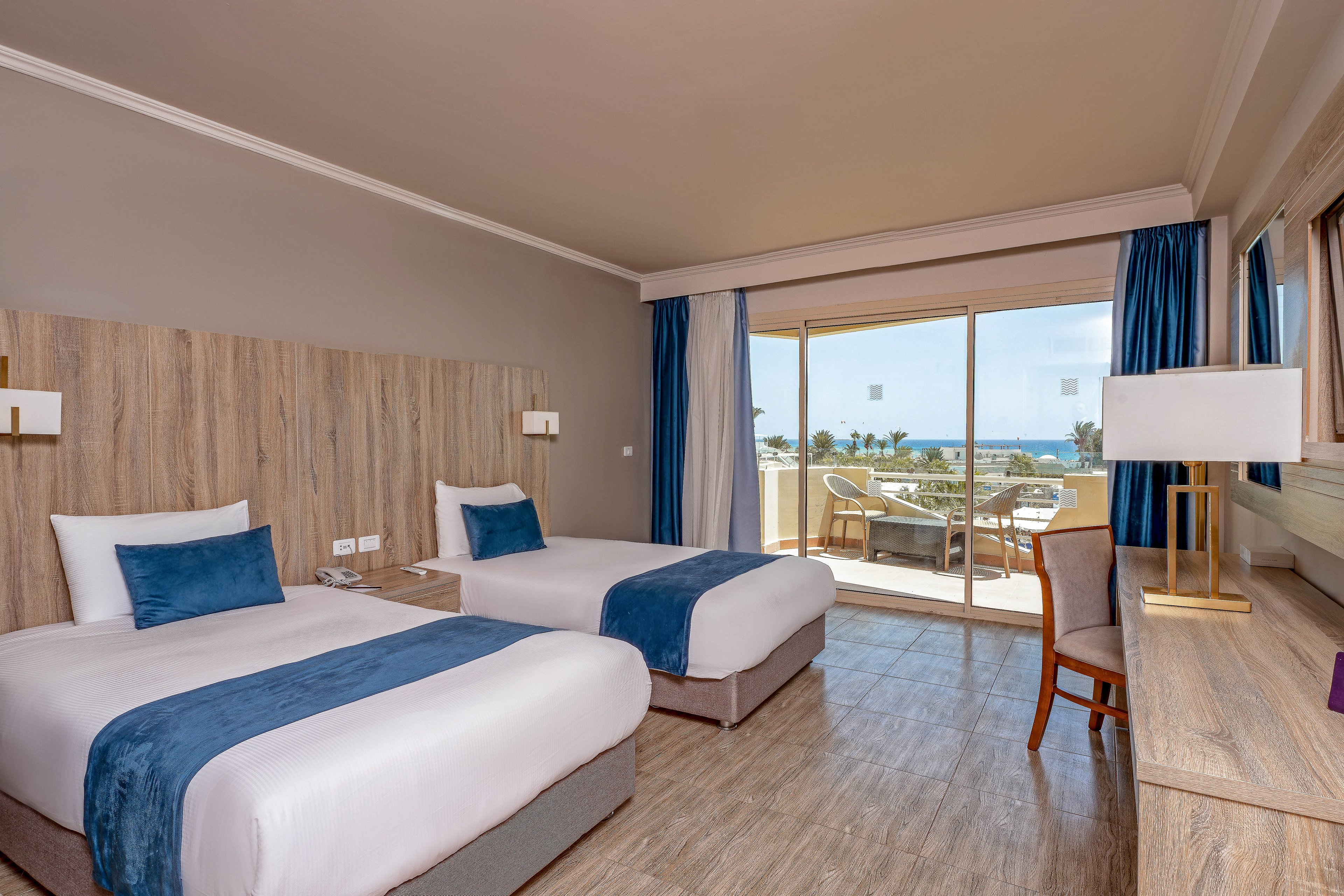 A contemporary hotel room features two neatly made twin beds with blue accents and a wooden headboard. The space is bright, with natural light streaming in from a large glass door leading to a balcony overlooking a scenic outdoor area. The room includes a desk, chair, and neutral-toned decor, creating a calm and inviting atmosphere. No visible text or numbers are present in the image.