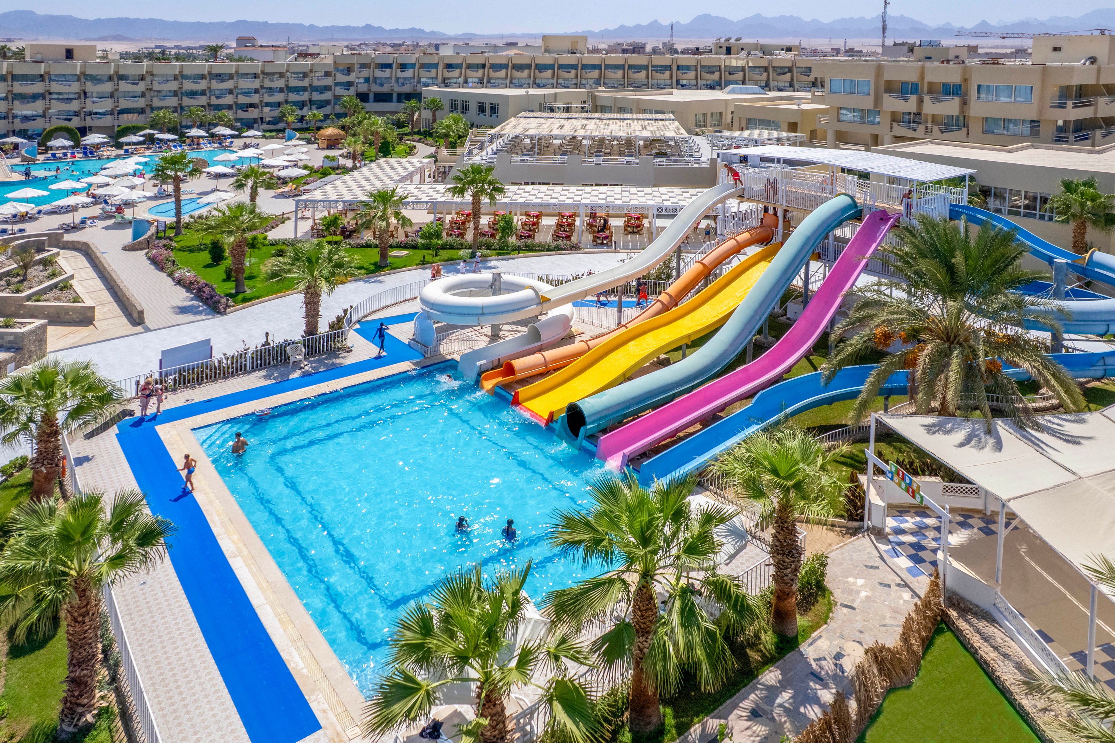 A large outdoor waterpark features multiple vibrant water slides leading into a spacious swimming pool. Several people are enjoying the pool, while others relax on sun loungers under umbrellas. The setting is a modern resort complex with palm trees, clear blue skies, and distant mountains, creating a lively and inviting atmosphere.