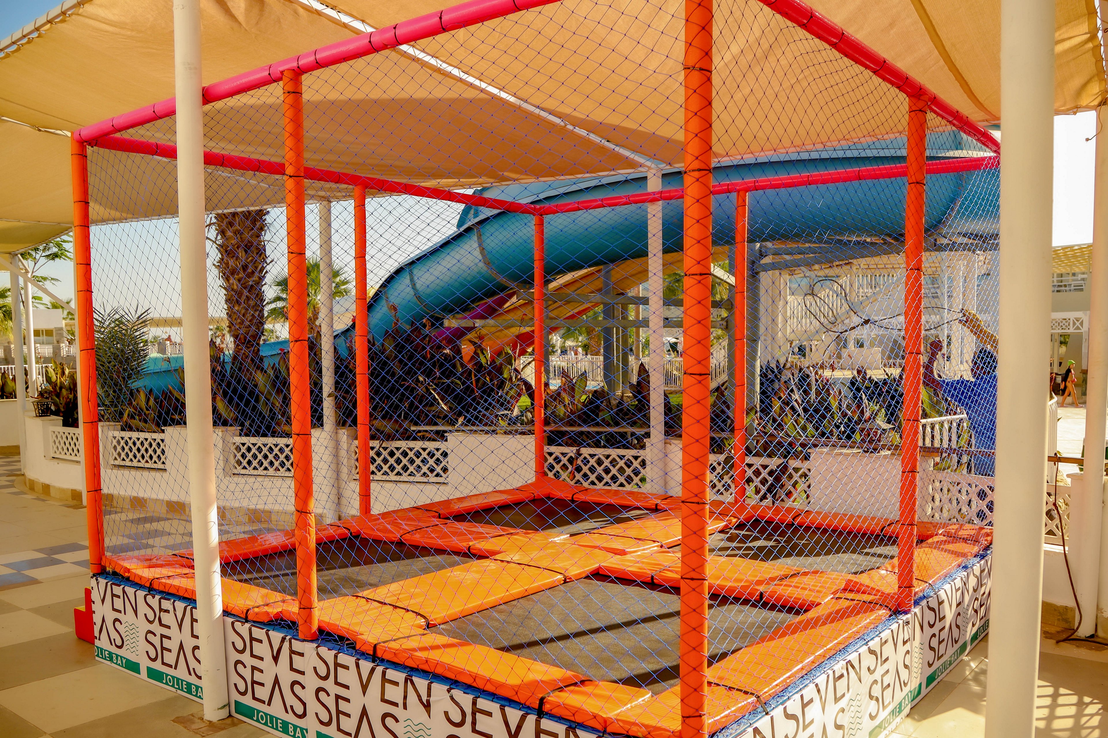 A brightly colored trampoline enclosure is set up outdoors under a canopy, surrounded by a safety net. The setting appears to be a recreational or water park area, with a large blue water slide visible in the background. The word 'SEVEN SEAS' is clearly visible on the side of the trampoline structure. The scene is sunlit, with palm trees and people in the background.