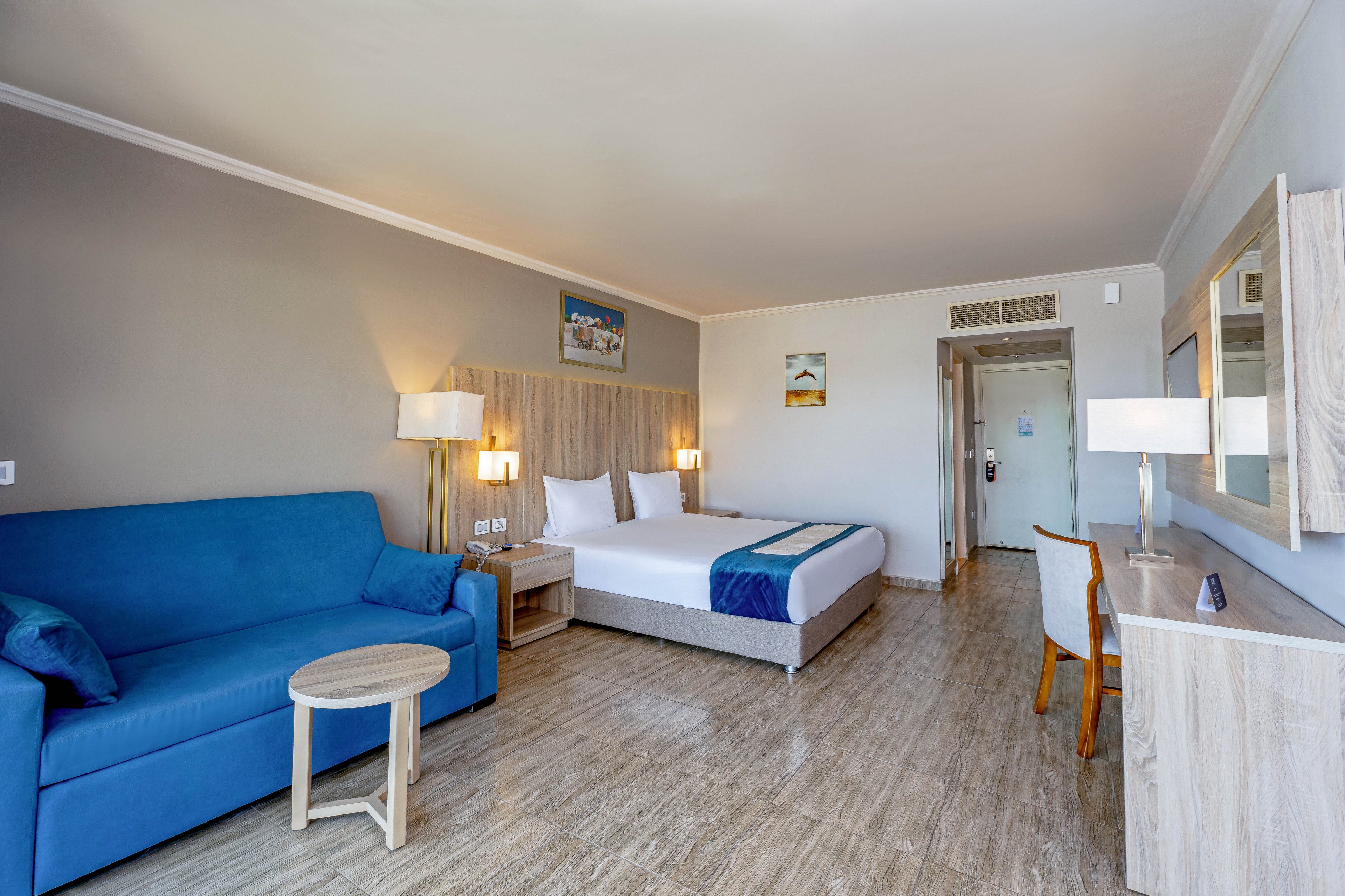 A spacious, contemporary hotel room features a double bed with white linens and a blue accent runner. The room includes a blue sofa, a wooden desk with a chair, and two bedside lamps, all set against neutral walls and wood-look flooring. Wall art and a large mirror add decorative touches, while natural light brightens the space. The setting is clean and inviting, ideal for relaxation or business travel.