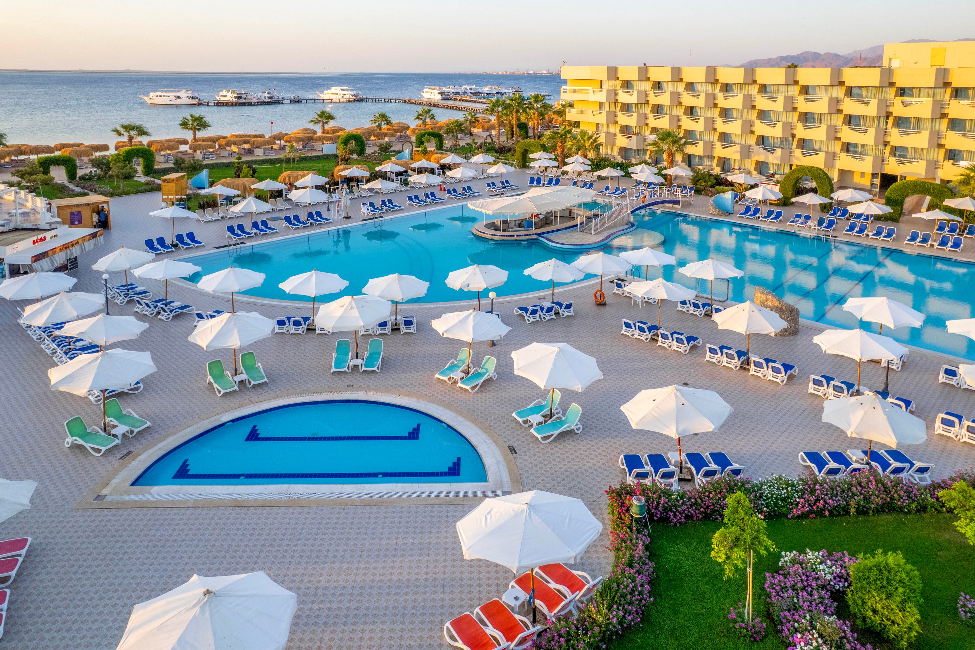 A spacious resort features multiple swimming pools surrounded by white sun umbrellas and lounge chairs. The setting overlooks the sea with several yachts docked in the distance, and a multi-story hotel building lines one side of the pool area. The scene is bright and inviting, with clear blue water and a relaxed vacation atmosphere. No visible text or numeric values are present in the image.