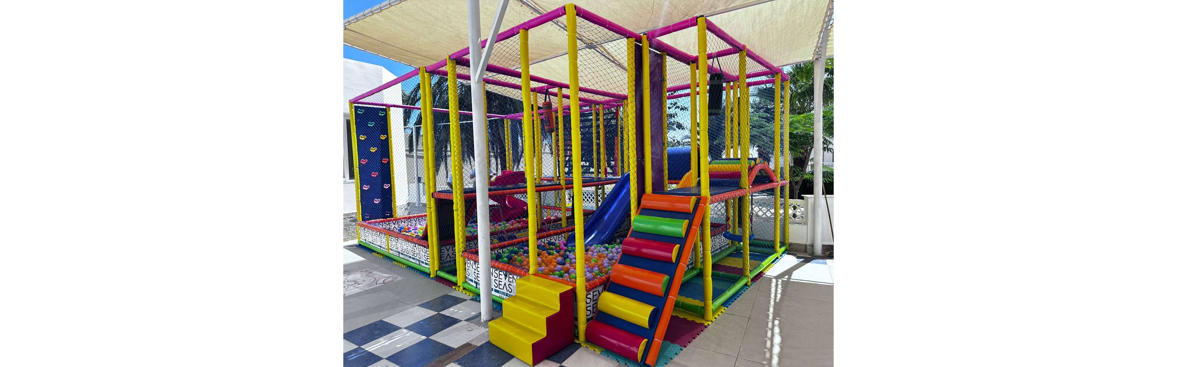 A vibrant, multi-level soft play structure is set outdoors under a canopy. The play area features a ball pit, climbing steps, slides, and padded barriers in bright colors. The flooring is checkered, and the structure is enclosed with safety netting. The setting is sunny with visible palm trees in the background. The words 'SEVEN SEAS' are clearly visible on the lower panels.