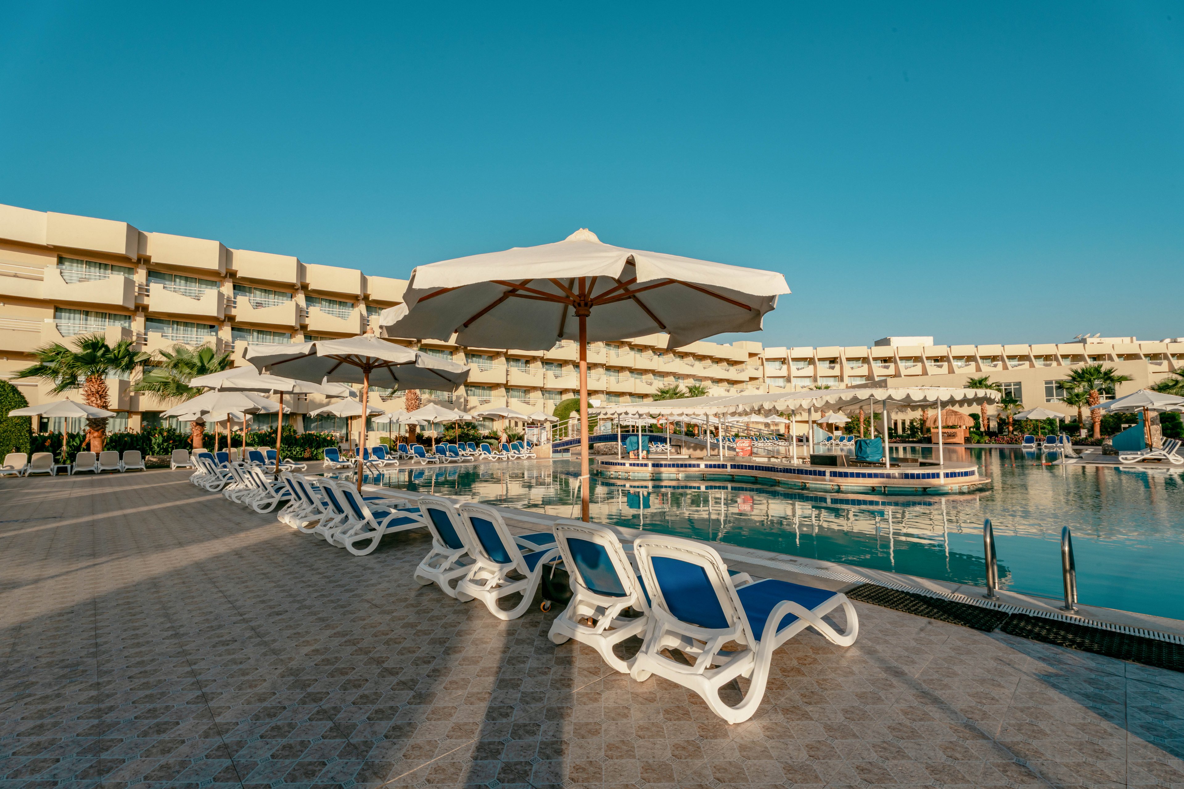 A spacious outdoor pool area at a modern resort features rows of white and blue lounge chairs under large white umbrellas. The pool is surrounded by a tiled deck and bordered by palm trees, with a multi-story hotel building in the background. The scene is brightly lit by clear, sunny weather, creating a relaxing and inviting atmosphere.