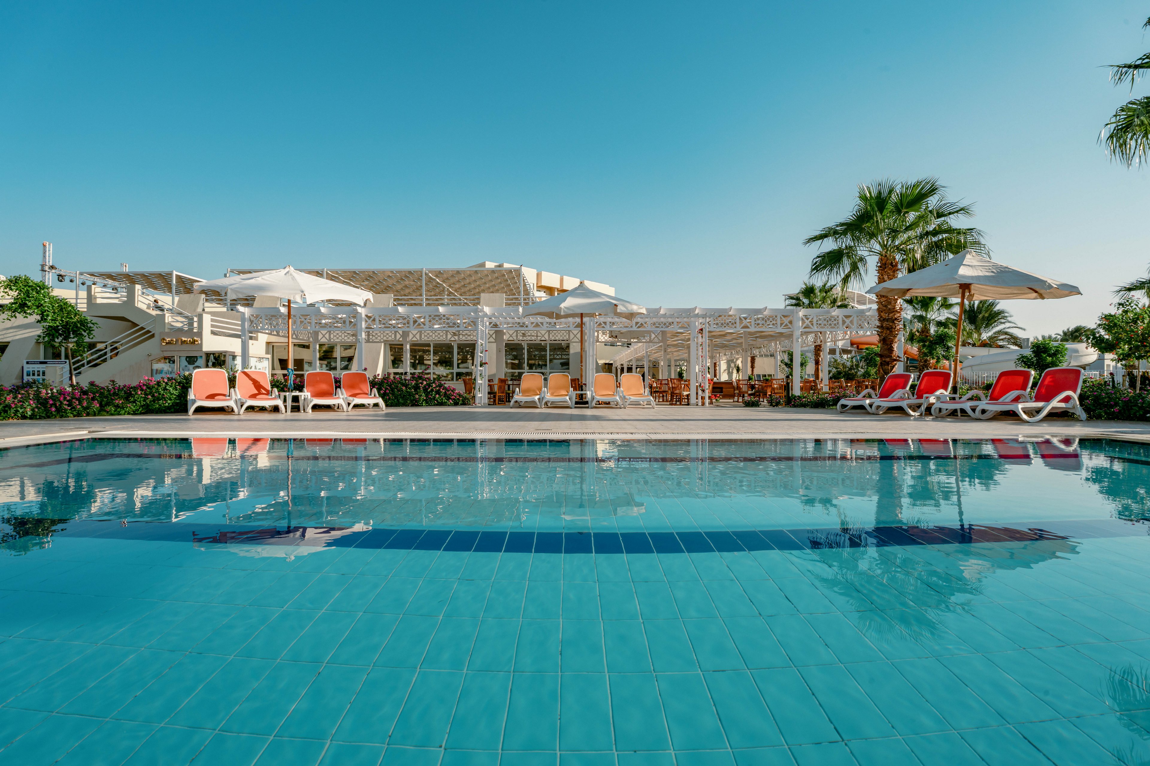 A tranquil outdoor swimming pool is the main focus, surrounded by orange and white lounge chairs and large white umbrellas. The setting is a modern resort with palm trees and clear blue skies, evoking a relaxing vacation atmosphere. The pool water is calm, reflecting the surrounding architecture and greenery.