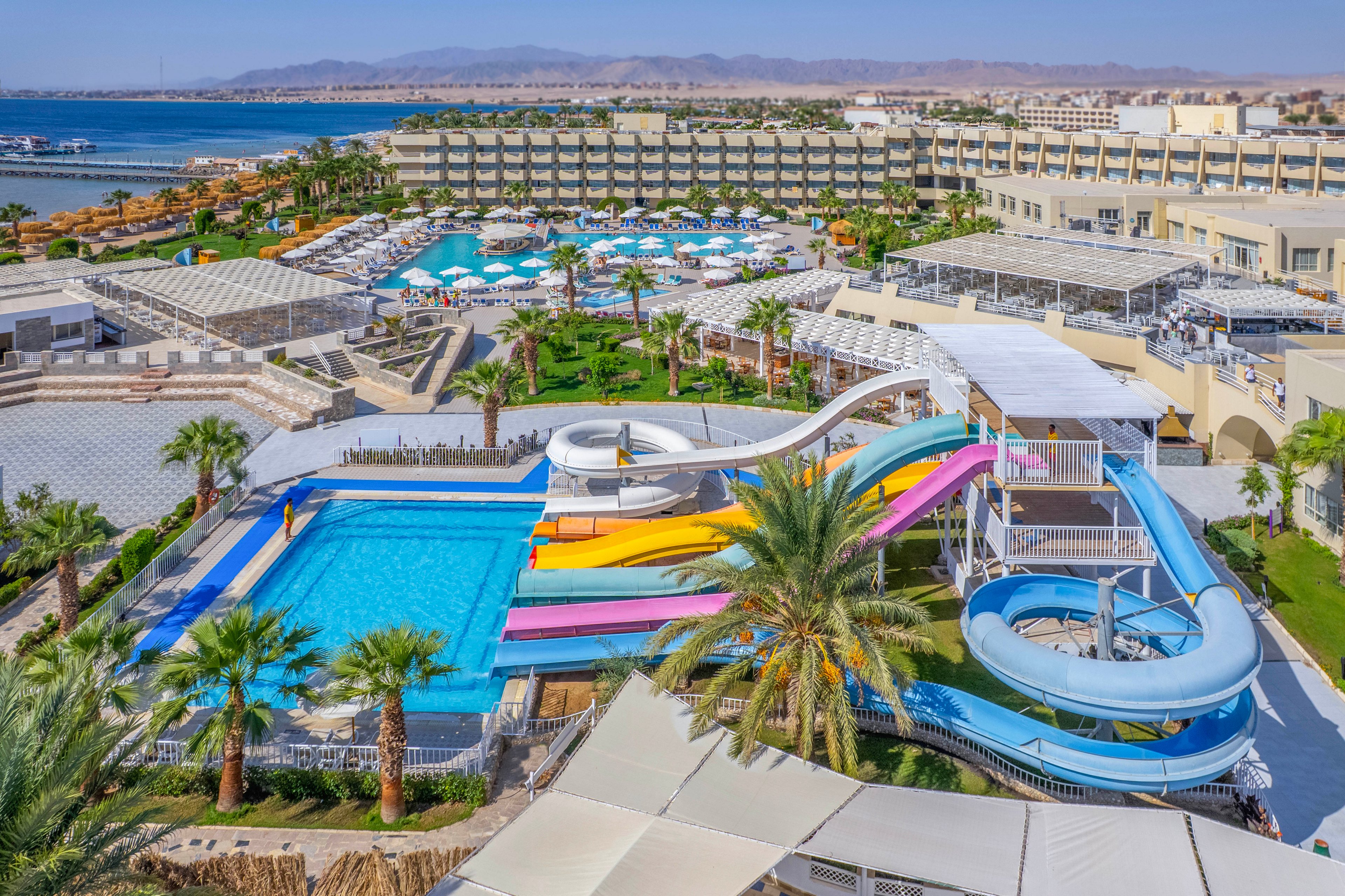 A large resort waterpark features multiple bright water slides leading into a spacious outdoor pool. The setting includes palm trees, sun loungers, and several pools surrounded by modern resort buildings. The scene is lively and inviting, with clear blue skies and distant mountains visible. The overall mood is vibrant and family-friendly, ideal for vacation and leisure activities.