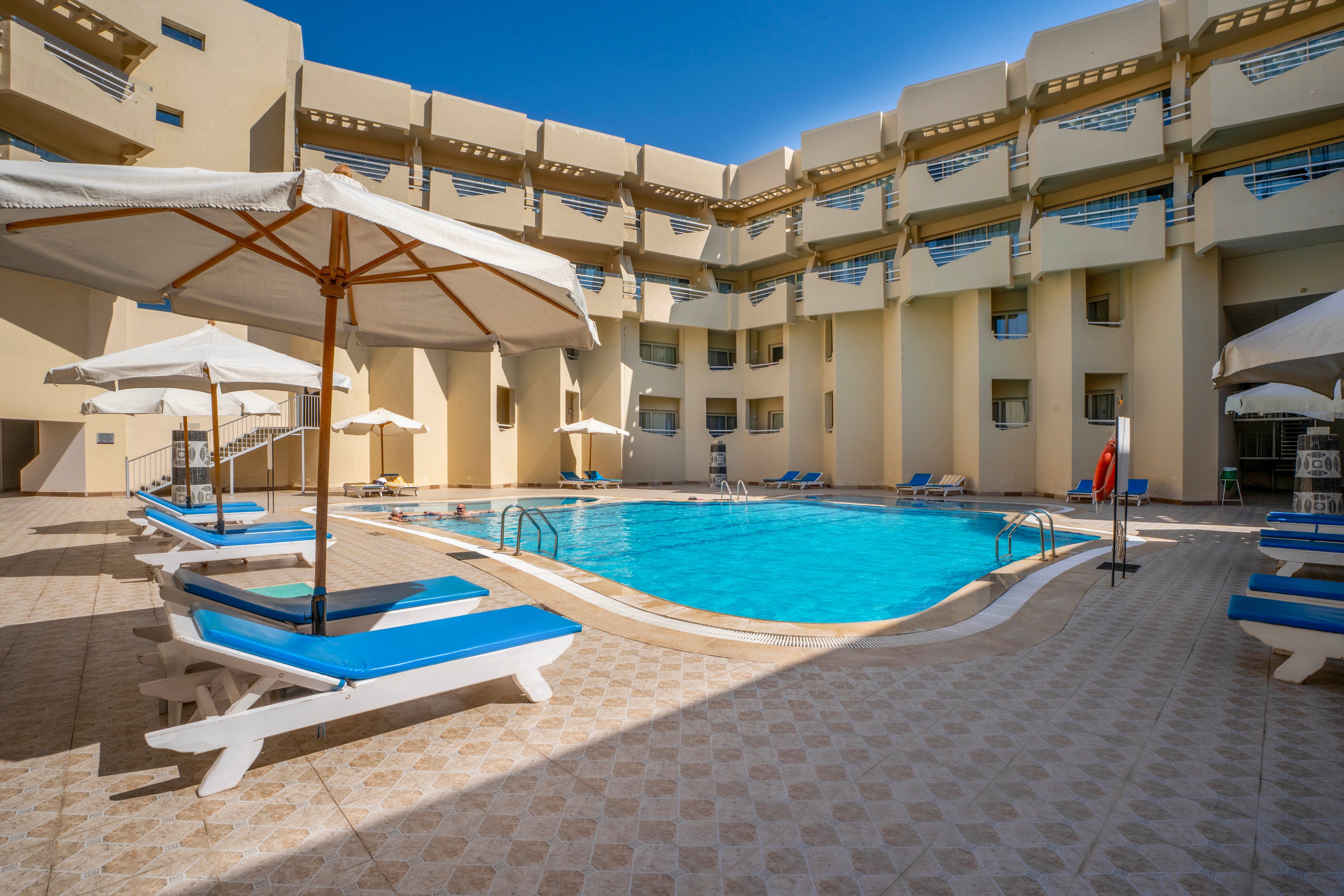 A bright hotel courtyard features a clear blue swimming pool surrounded by white lounge chairs and large umbrellas. The modern beige building encloses the pool area, creating a private and relaxing atmosphere. The tiled pool deck is sunlit, and the scene is empty of people, emphasizing tranquility and leisure. No visible text or numbers are present in the image.