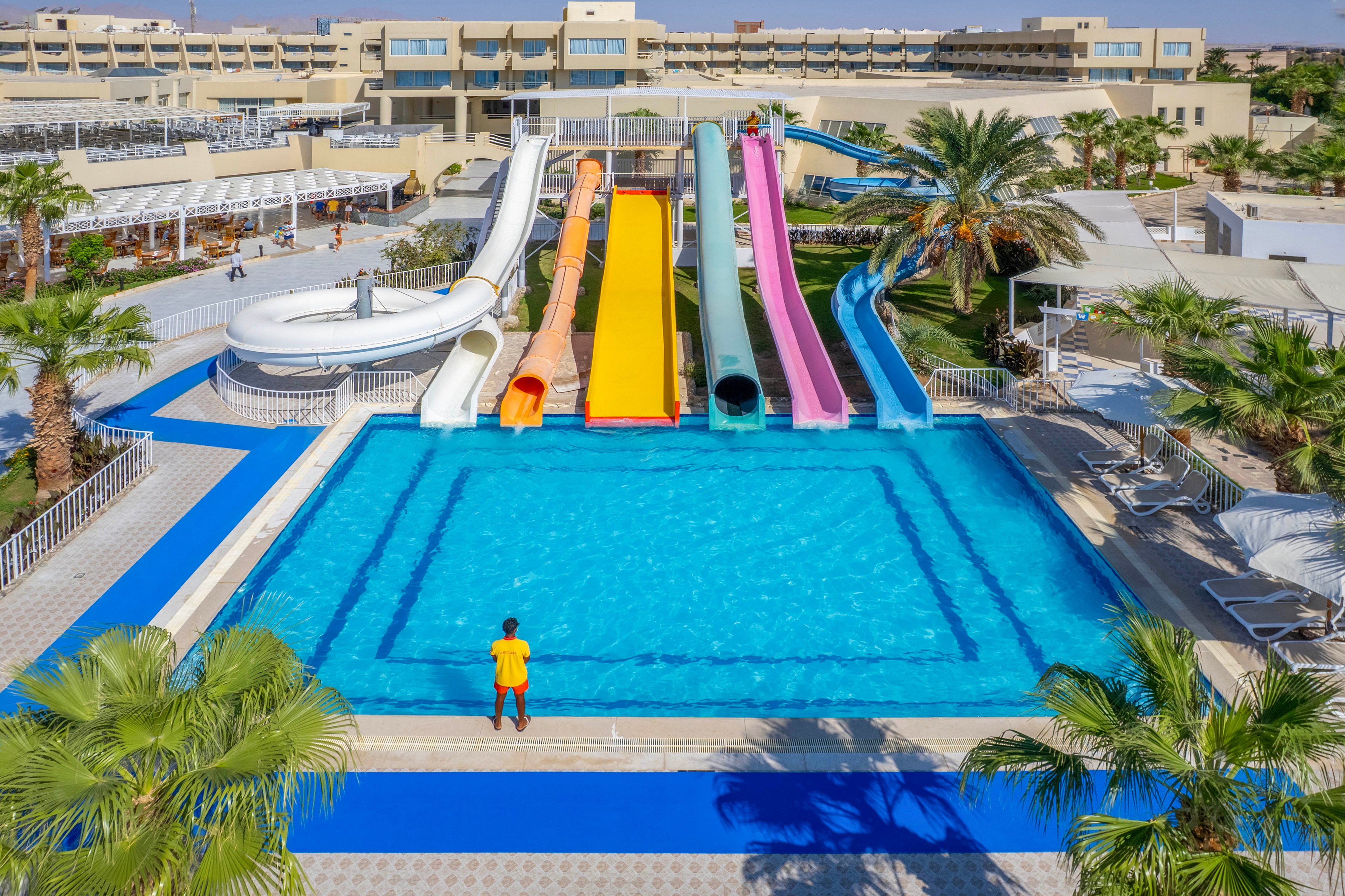 A large outdoor swimming pool features multiple colorful water slides, including white, yellow, orange, red, blue, and pink chutes. A single person in a yellow shirt stands at the pool's edge, facing the slides. The setting is a sunny resort with palm trees, lounge chairs, and modern buildings in the background, creating a vibrant and inviting atmosphere.