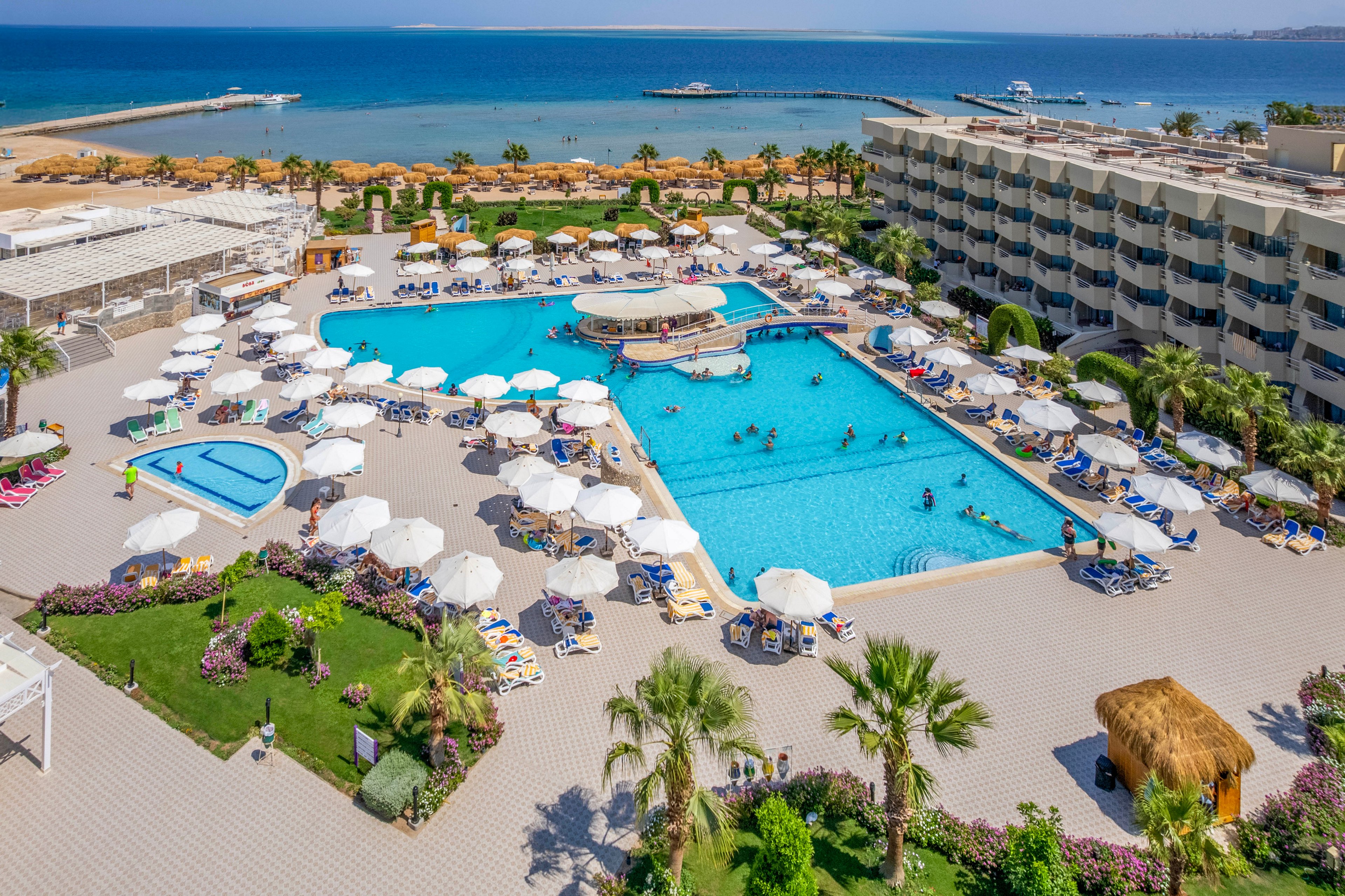 A large resort swimming pool is surrounded by white umbrellas, lounge chairs, and palm trees, with a smaller pool nearby. The scene overlooks a sandy beach and calm blue sea, with a pier and boats visible in the distance. Guests of various ages are enjoying the pool and sunbathing, creating a relaxed vacation atmosphere under clear skies.